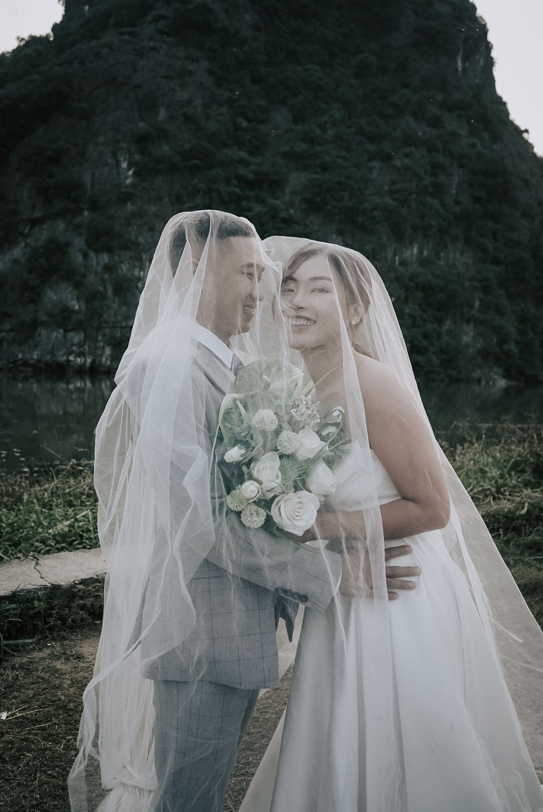 Romantic couple sharing a tender moment under a bridal veil with lush green mountains behind them.