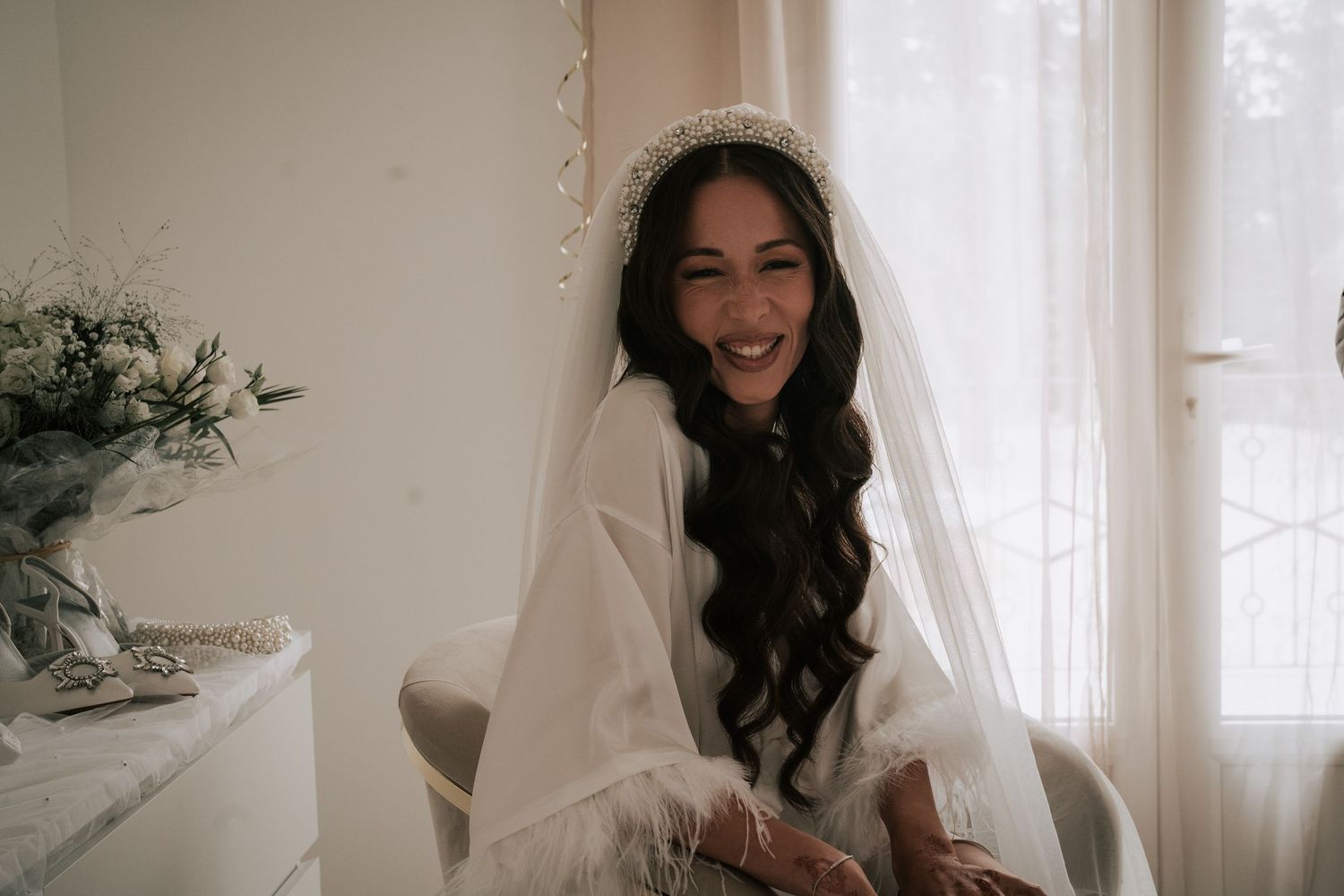 Smiling bride in white robe and veil with floral headpiece sitting by a bright window on her wedding day.