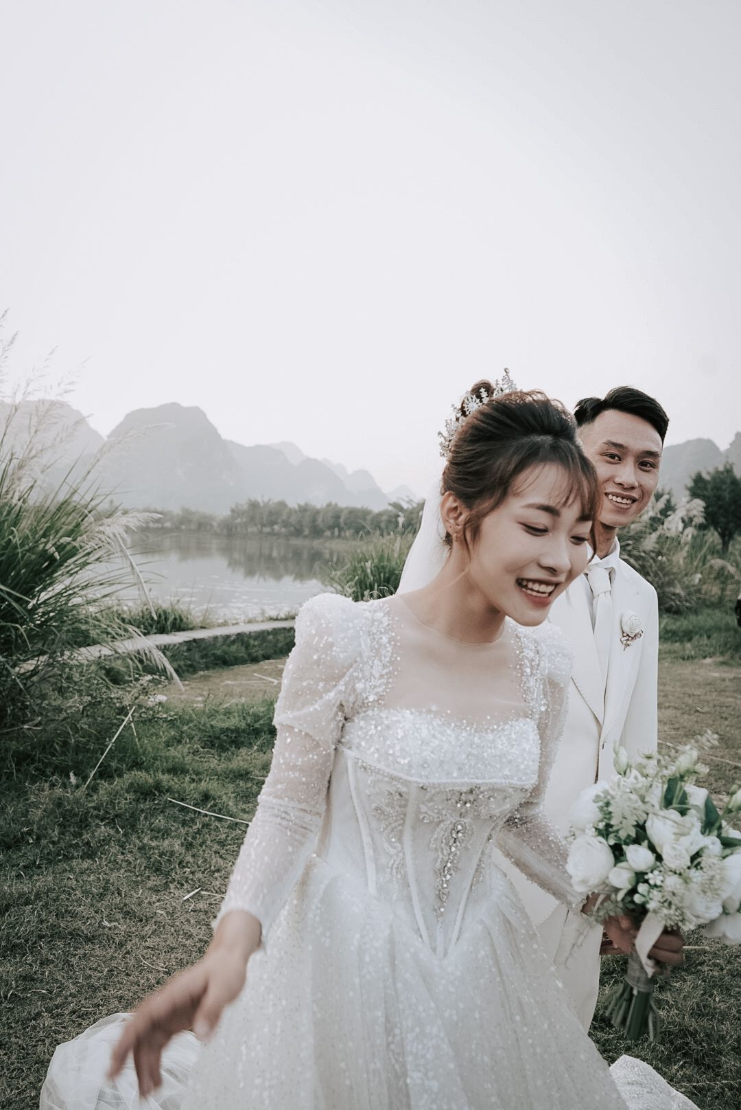 A smiling bride in a sparkling white gown holds a bouquet as a groom in white stands behind her near a scenic lake.