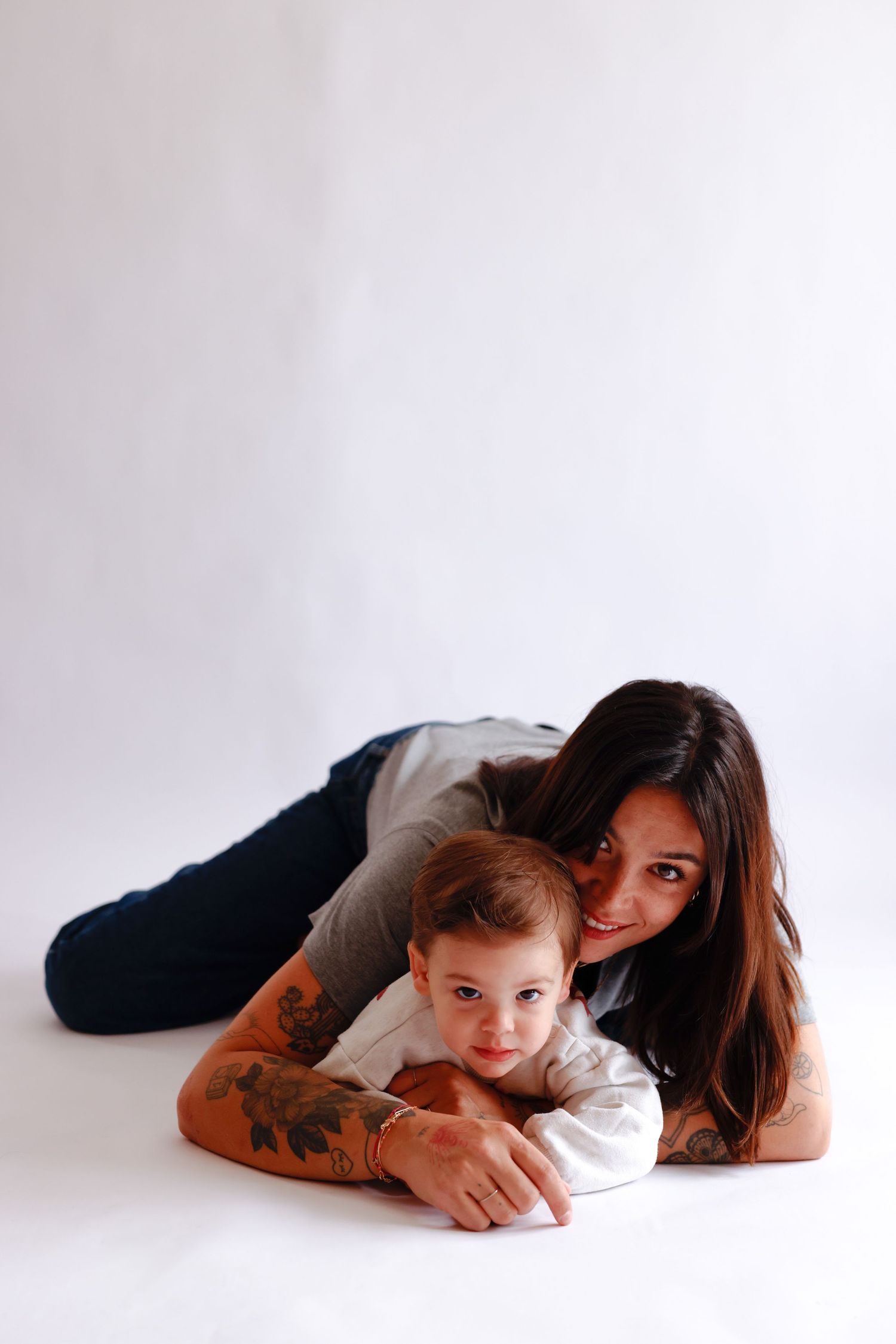 Tattooed mother lying on white floor smiling while embracing her baby boy.