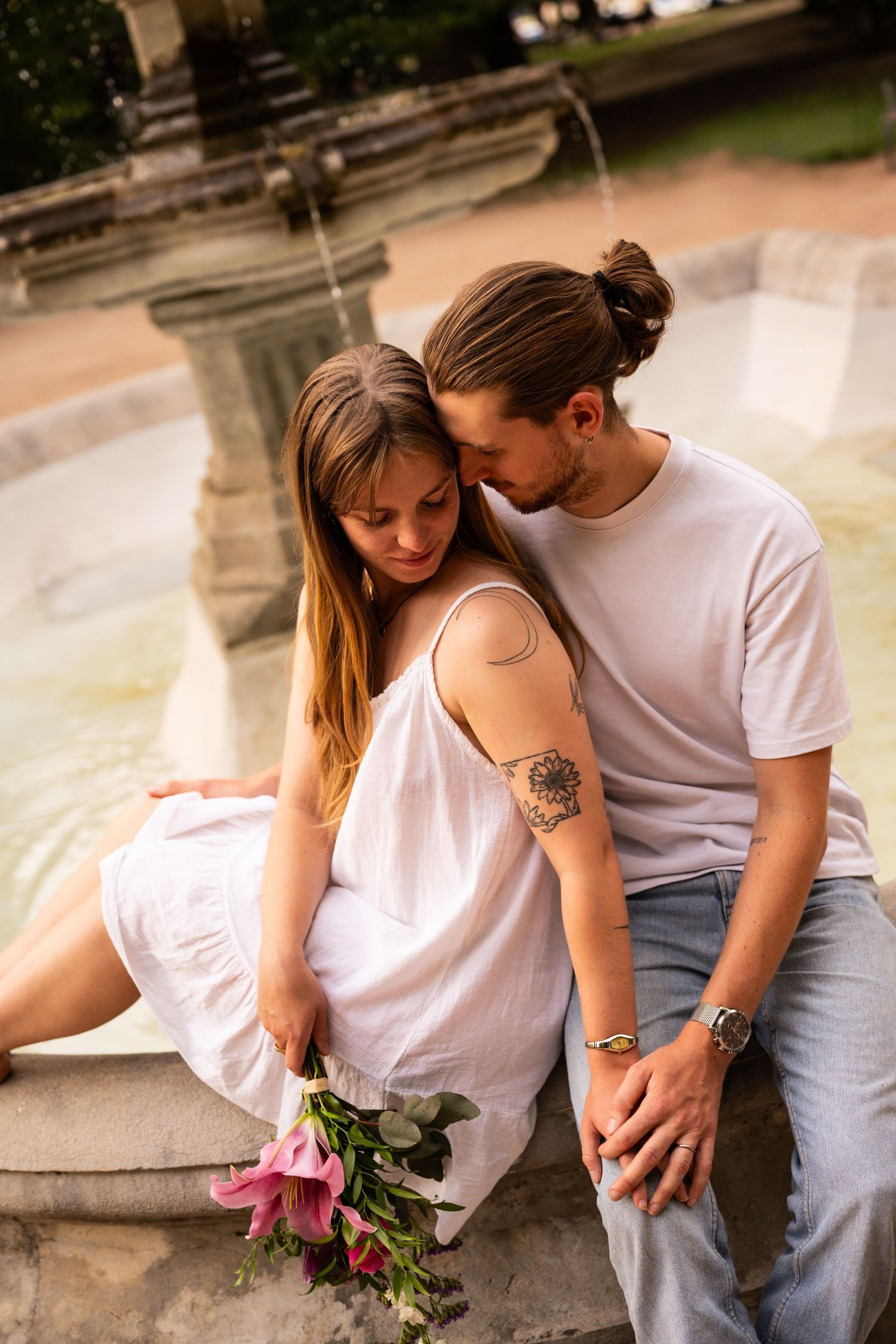 Couple sitting intimately by a stone fountain, man kissing womans head as she holds flowers.