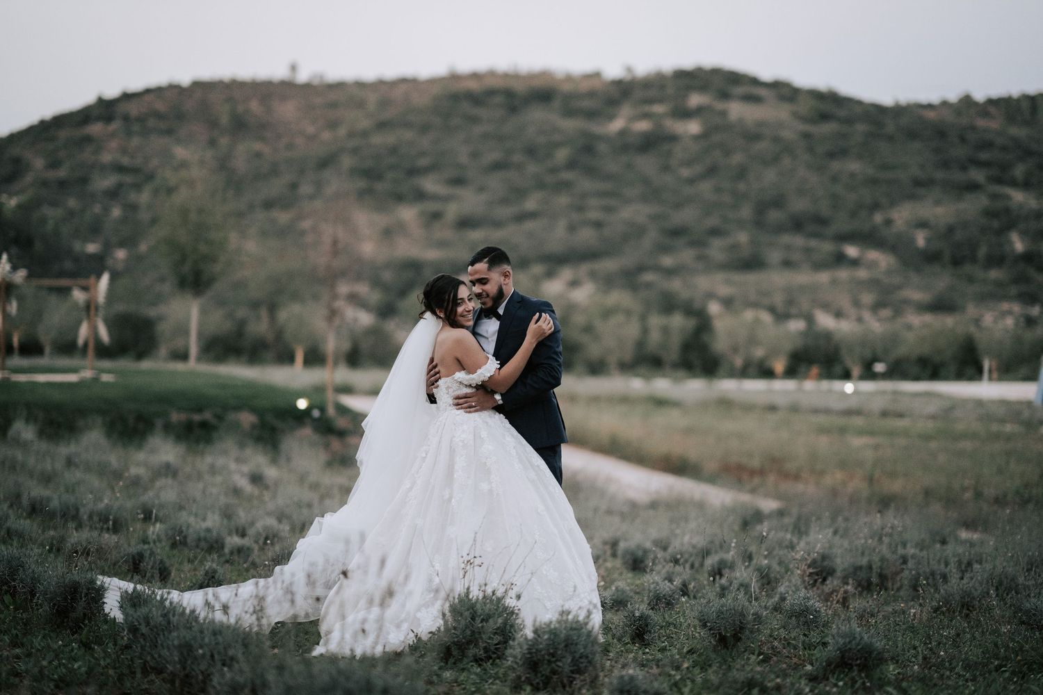 Bride in a flowing white gown and groom share a romantic embrace in a scenic outdoor field at dusk.