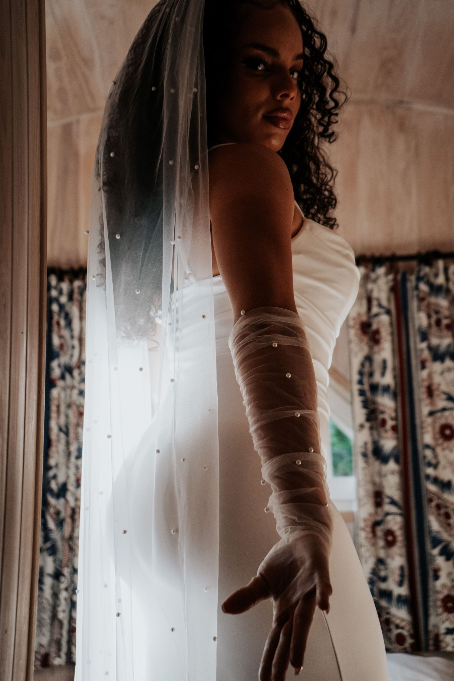 Bride in sleek white dress and pearl-embellished veil and gloves poses dramatically by a sunlit window.