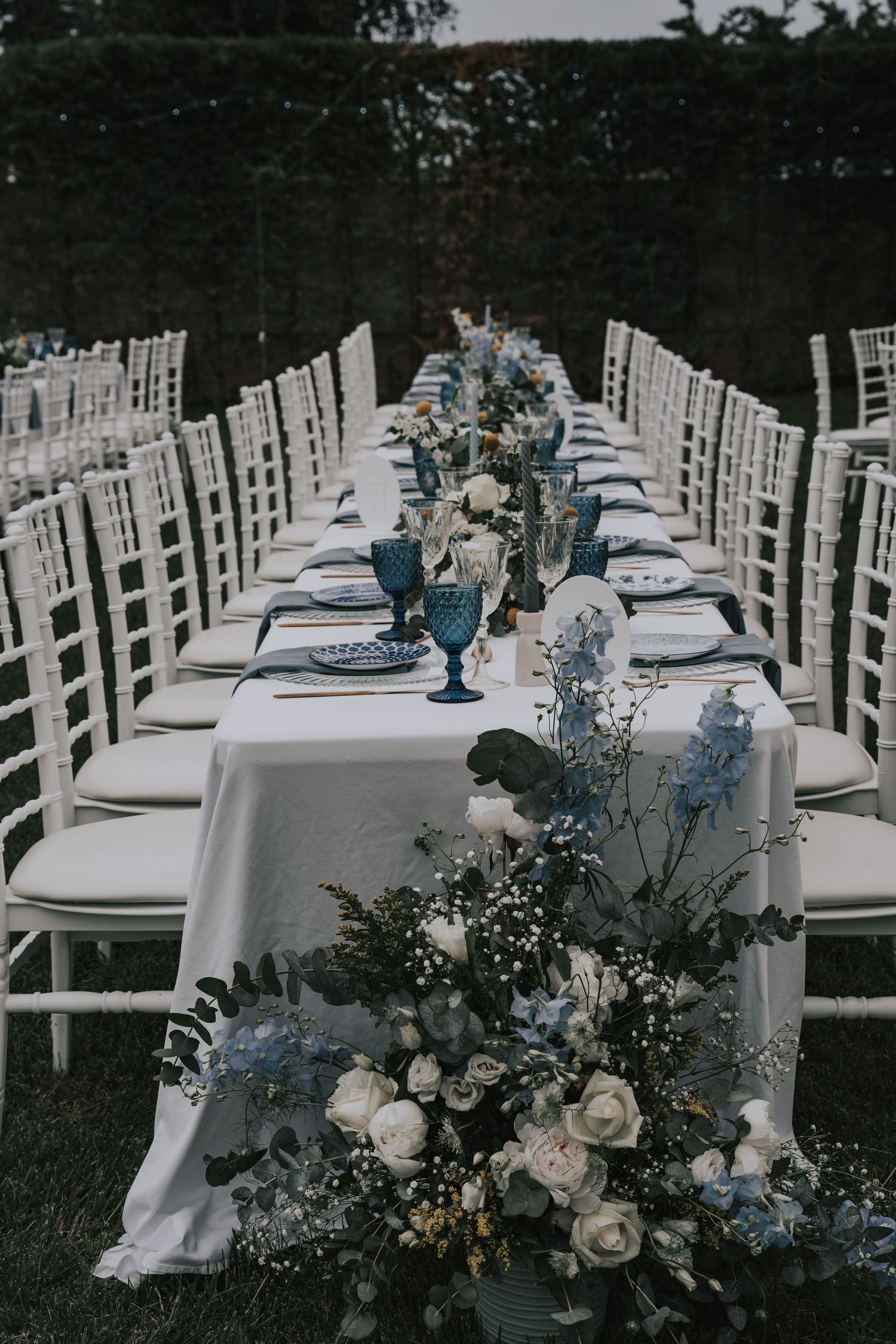Elegant outdoor wedding reception table with blue glassware, white florals, and chiavari chairs.
