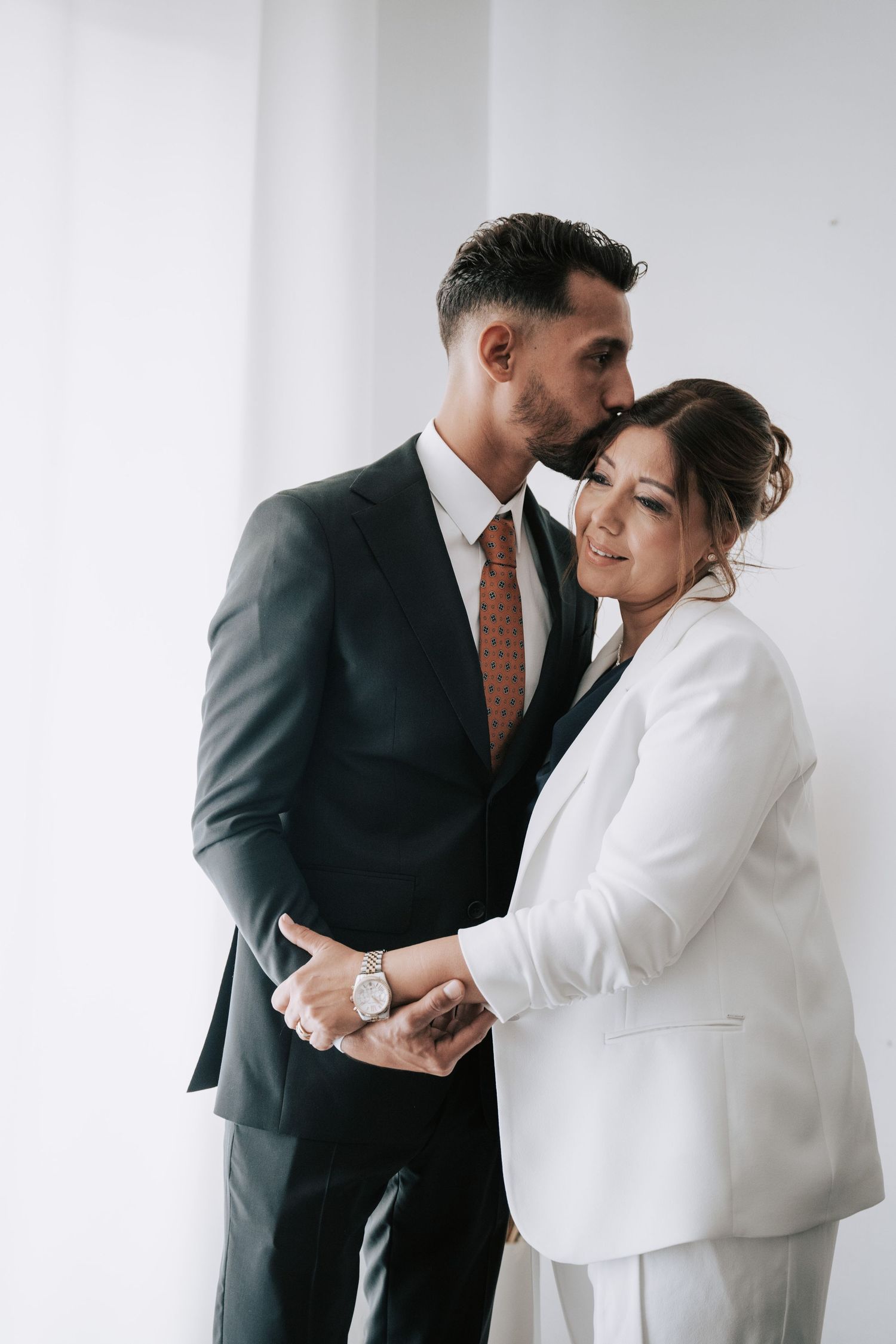 Groom in dark suit kissing bride in white blazer on the forehead in a tender moment.
