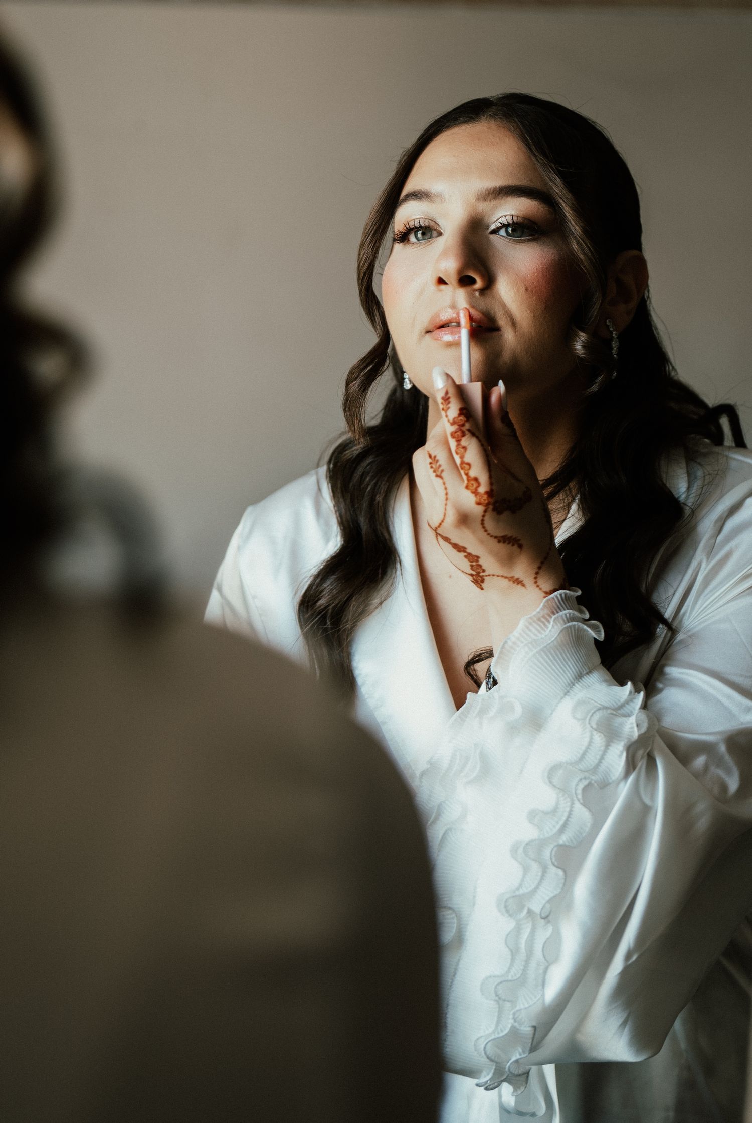 Beautiful bride in white robe applying lip gloss with henna-decorated hand while getting ready for her wedding.