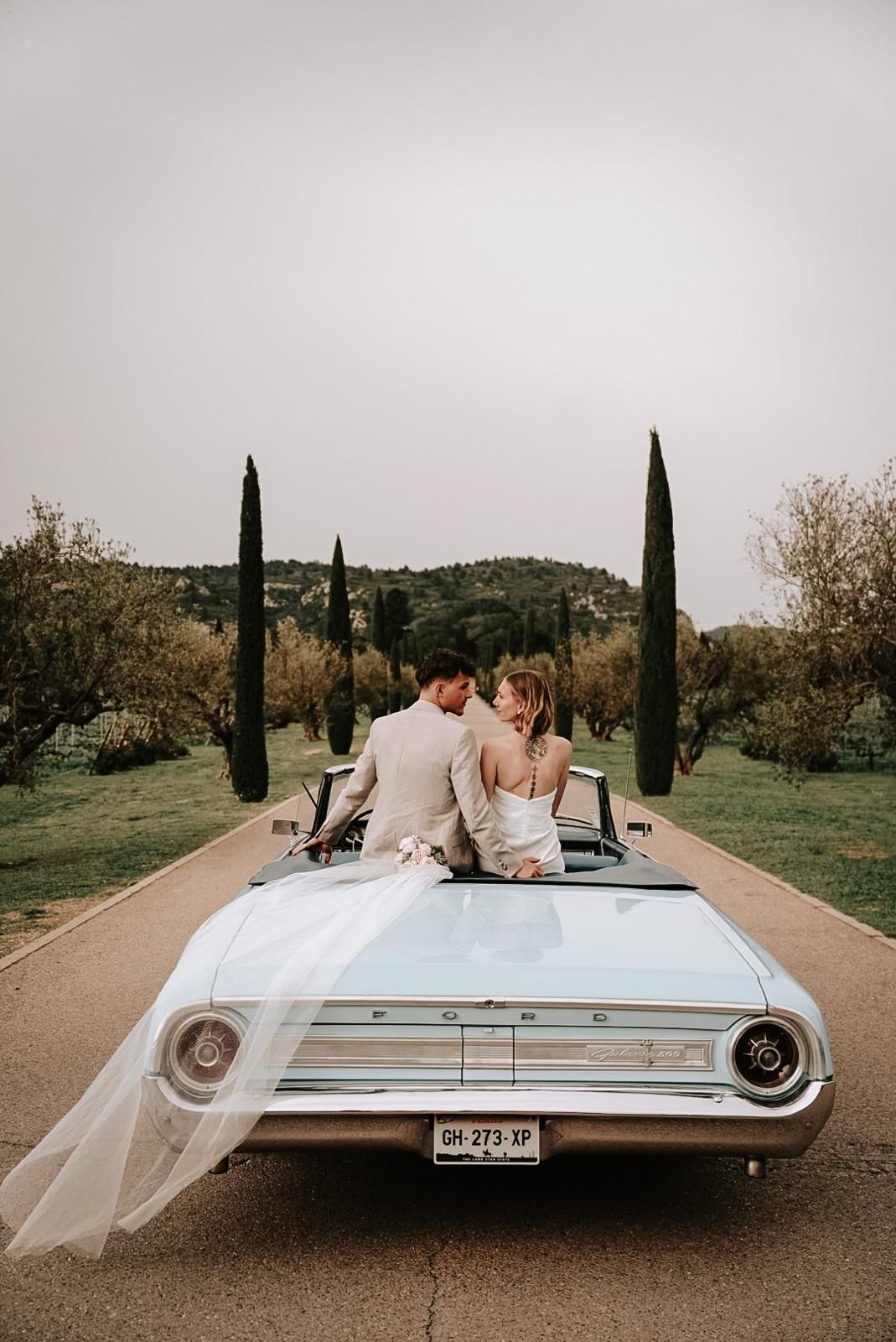 Bride and groom kiss on a white vintage Ford convertible, veil flowing, framed by tall cypress trees.