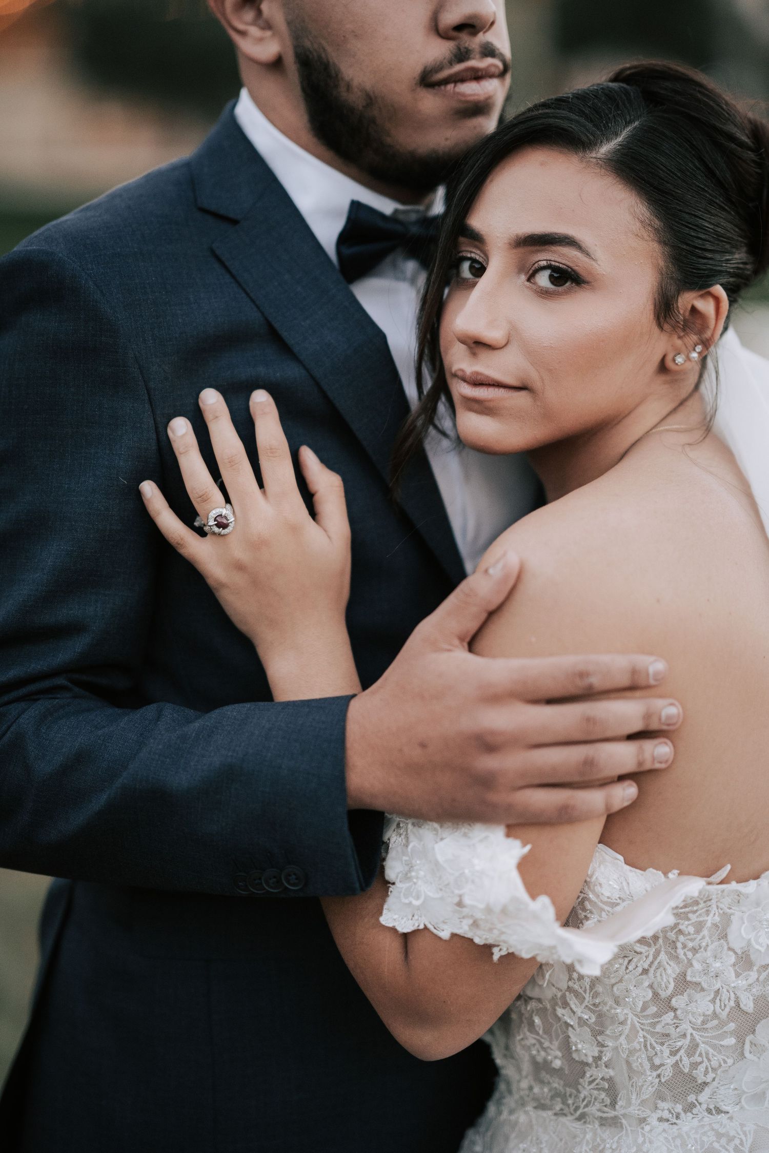 Close-up of bride leaning against groom in navy suit, displaying her floral ring and lace off-shoulder gown.