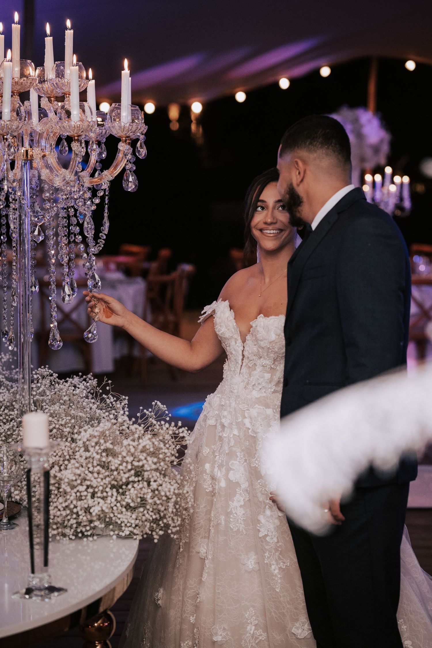 Bride in lace gown smiling with groom beside crystal candelabra centerpiece at elegant evening wedding reception.