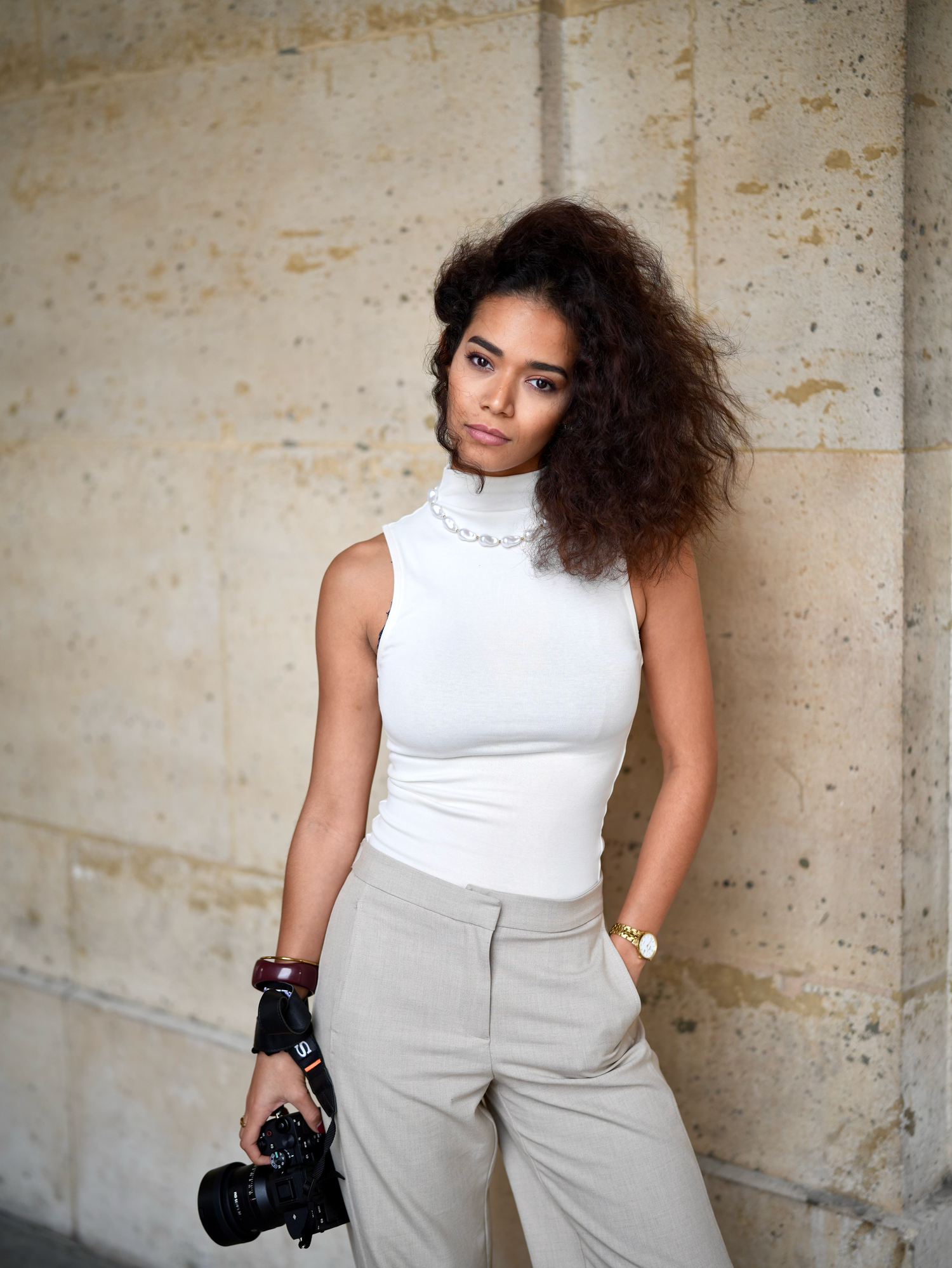 Young woman with curly hair in white sleeveless turtleneck and beige trousers holding a camera against a stone wall.