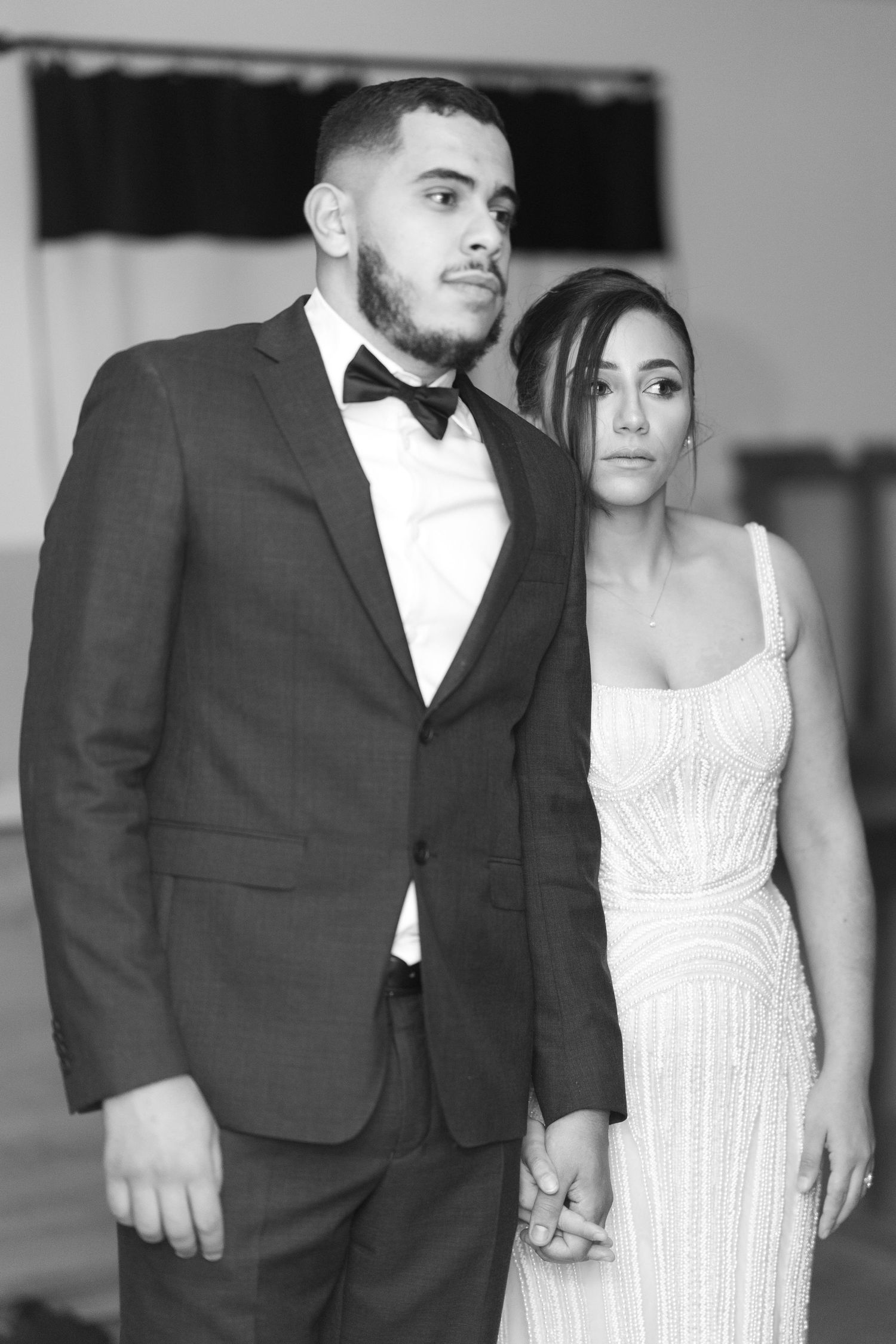 Elegant black and white portrait of bride and groom holding hands, groom in suit with bow tie, bride in beaded gown.