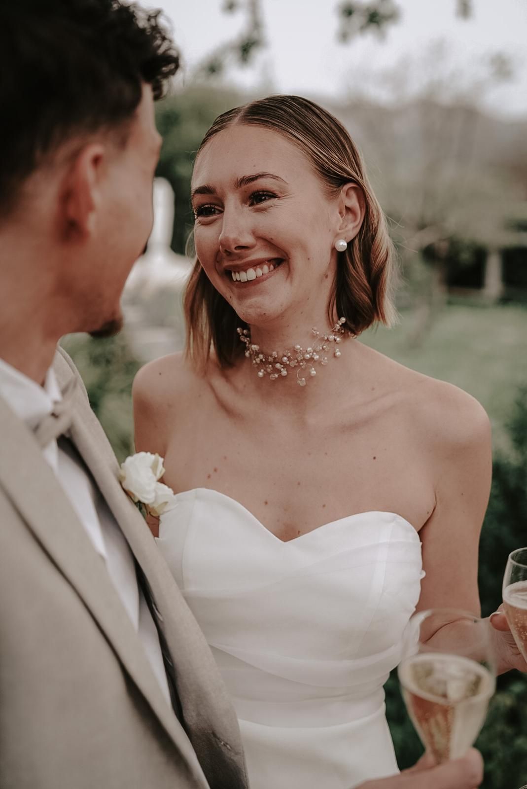 Radiant bride in strapless white gown and floral necklace smiles joyfully while holding a champagne glass.