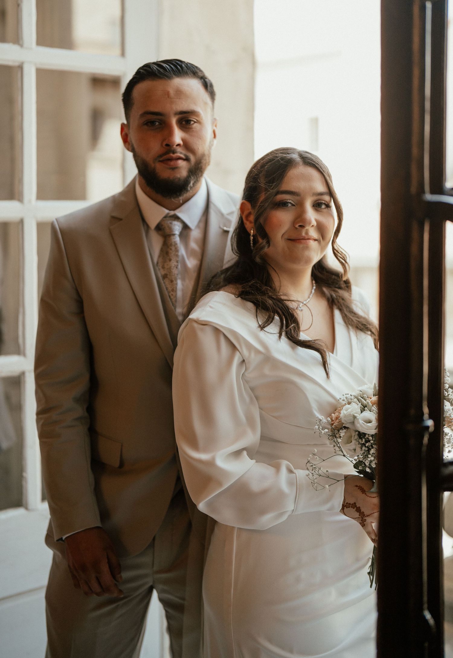 Bride and groom posing together in wedding attire, she in white dress holding bouquet, he in beige suit.