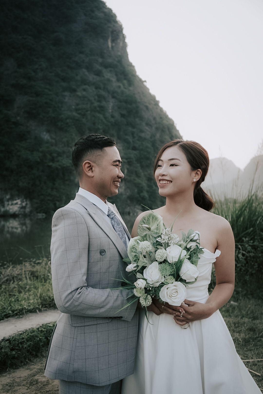 Smiling Asian couple in wedding attire pose with white bouquet against mountain backdrop.