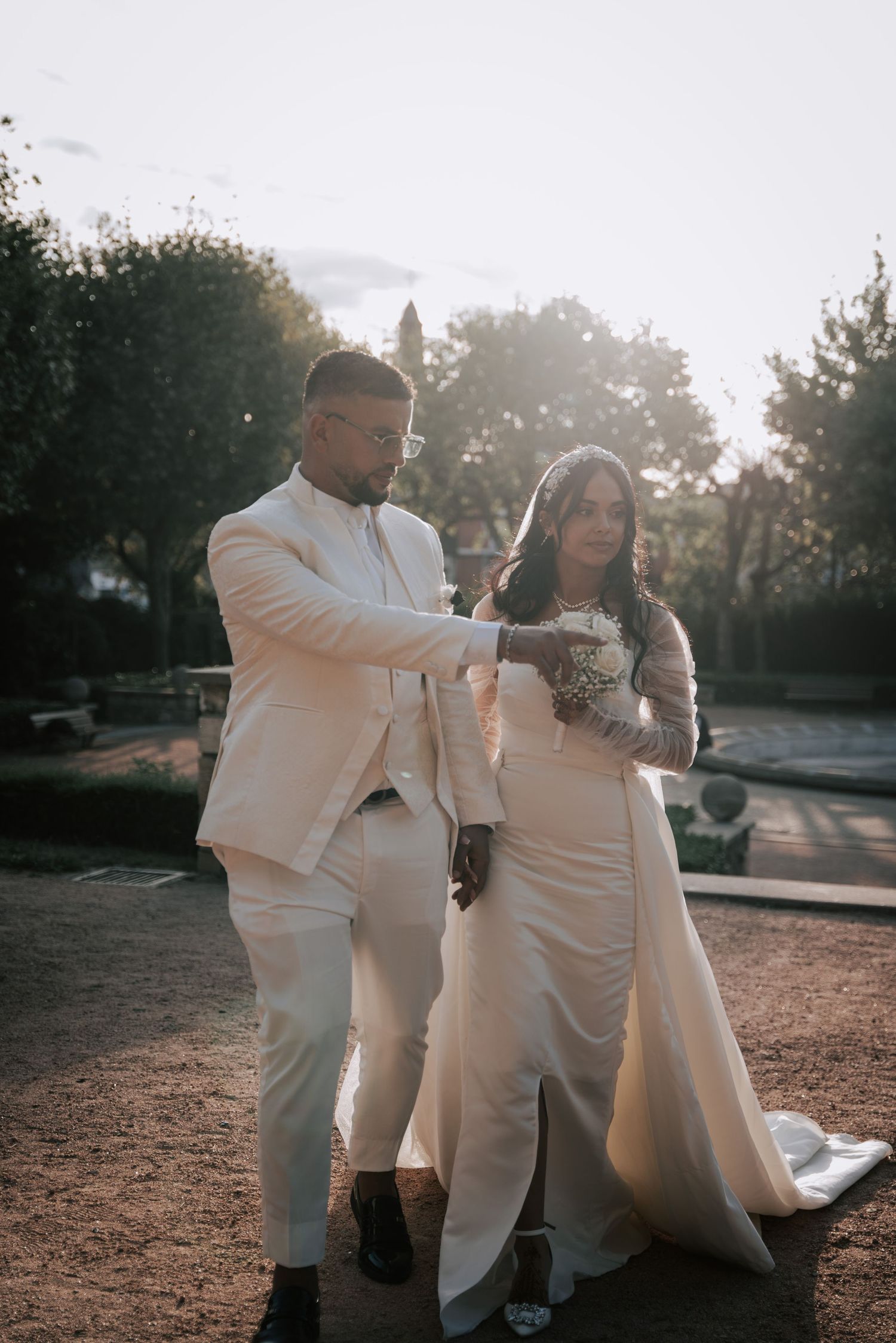 Bride and groom in matching white attire walk together at golden hour in an outdoor garden setting.