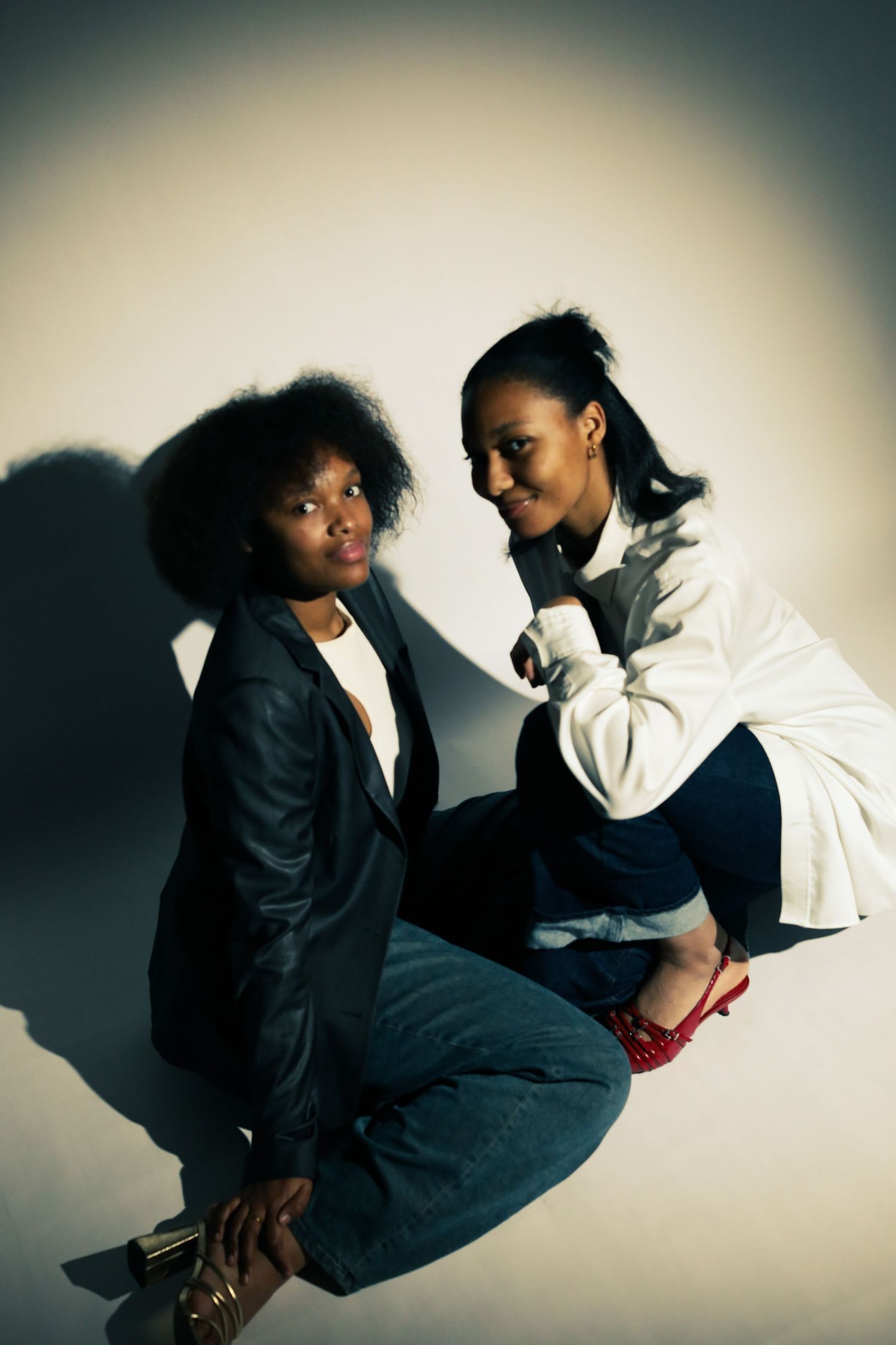 Two stylish Black women posing together, one in a black blazer with an afro, the other in a white jacket and red heels.