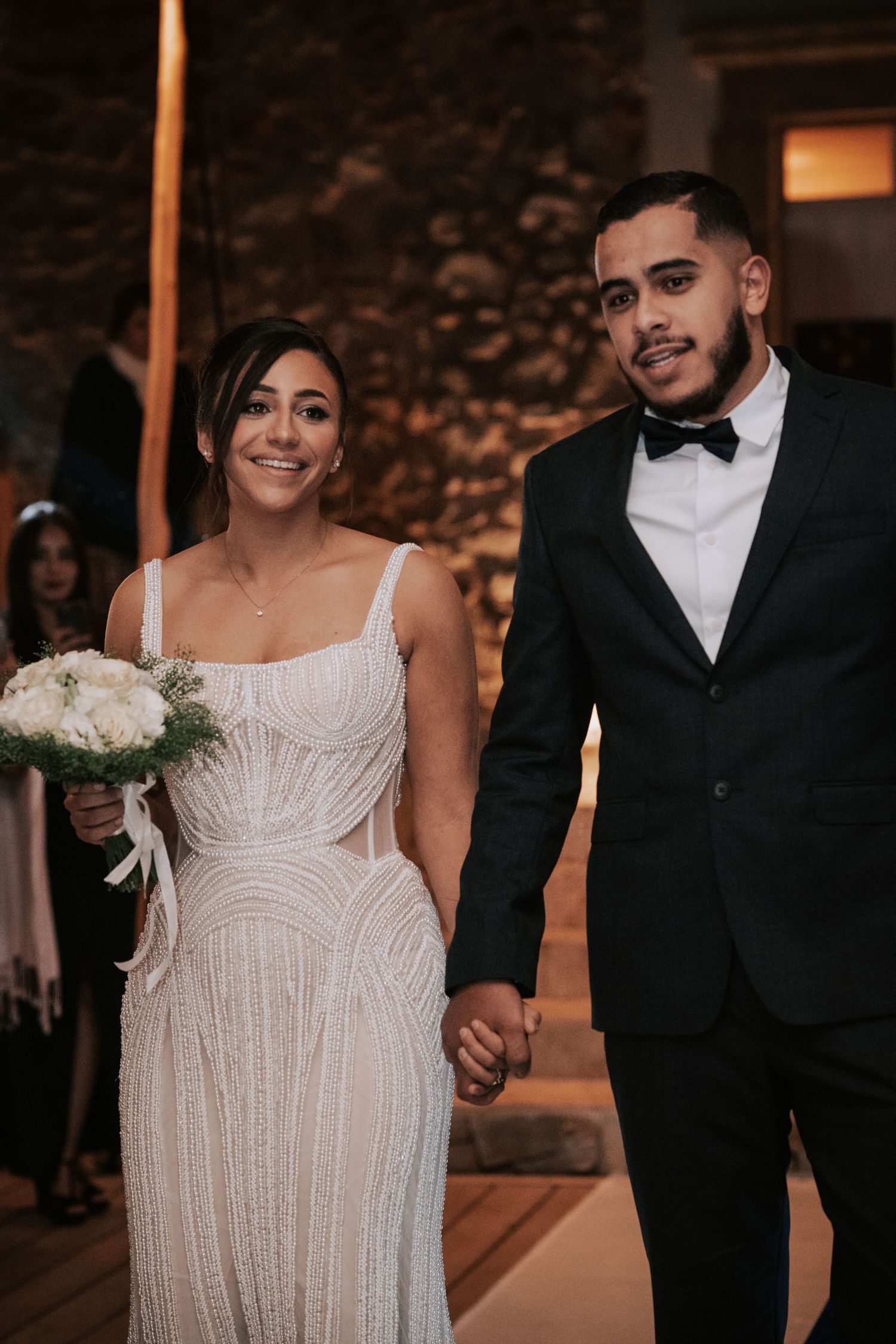 Smiling bride in beaded gown holding white bouquet walks hand in hand with groom after wedding ceremony.