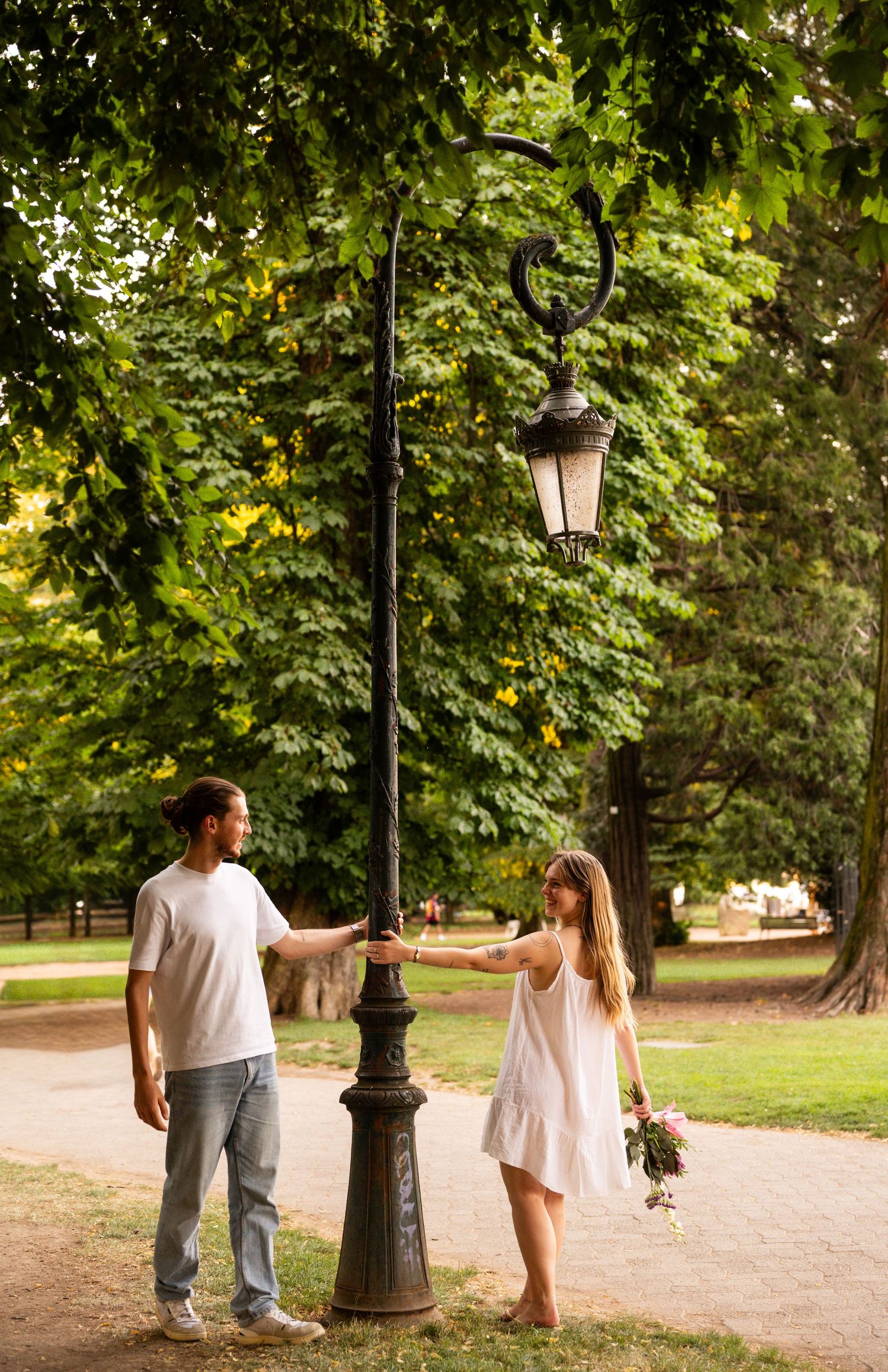 Couple holding hands around a vintage iron lamppost in a tree-lined park pathway.
