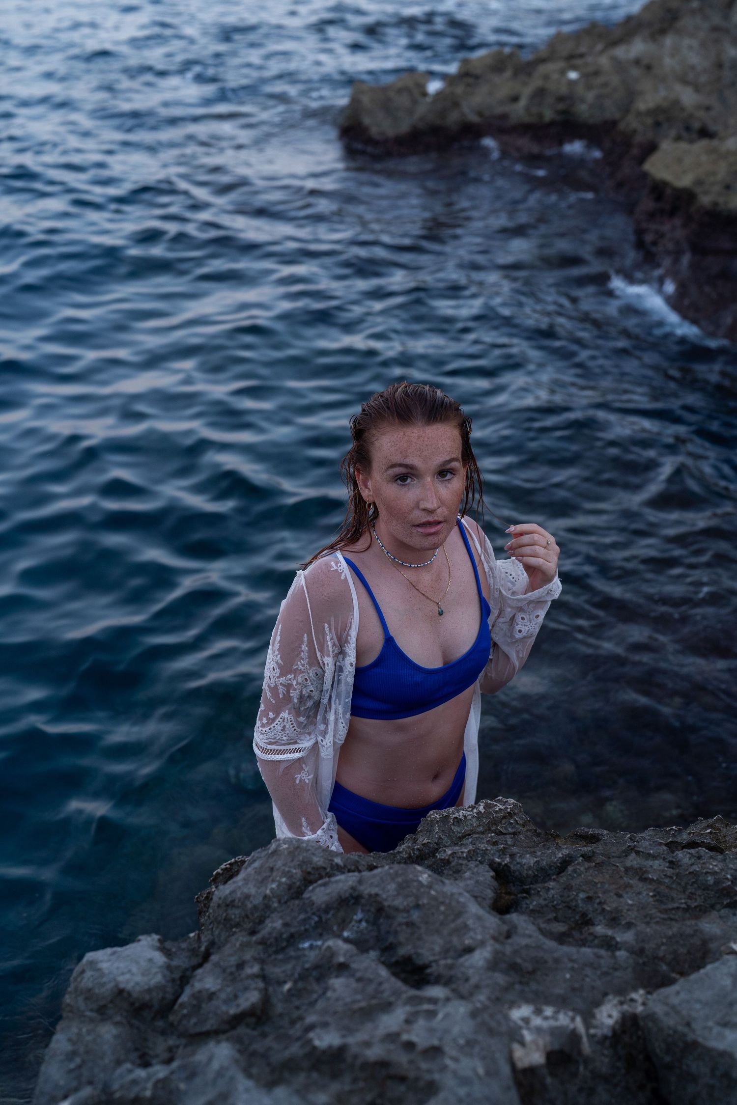 Woman in blue two-piece swimsuit and white lace cover-up posing on rocky ocean shore at dusk.
