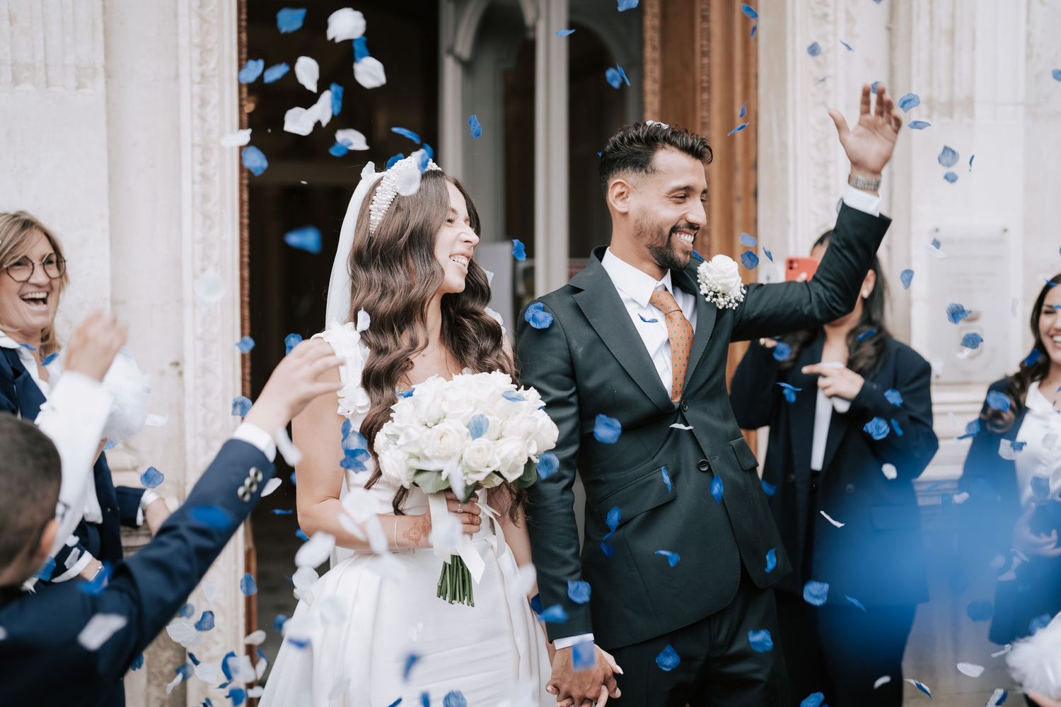 Joyful bride and groom exit church amid blue confetti with celebrating wedding guests.
