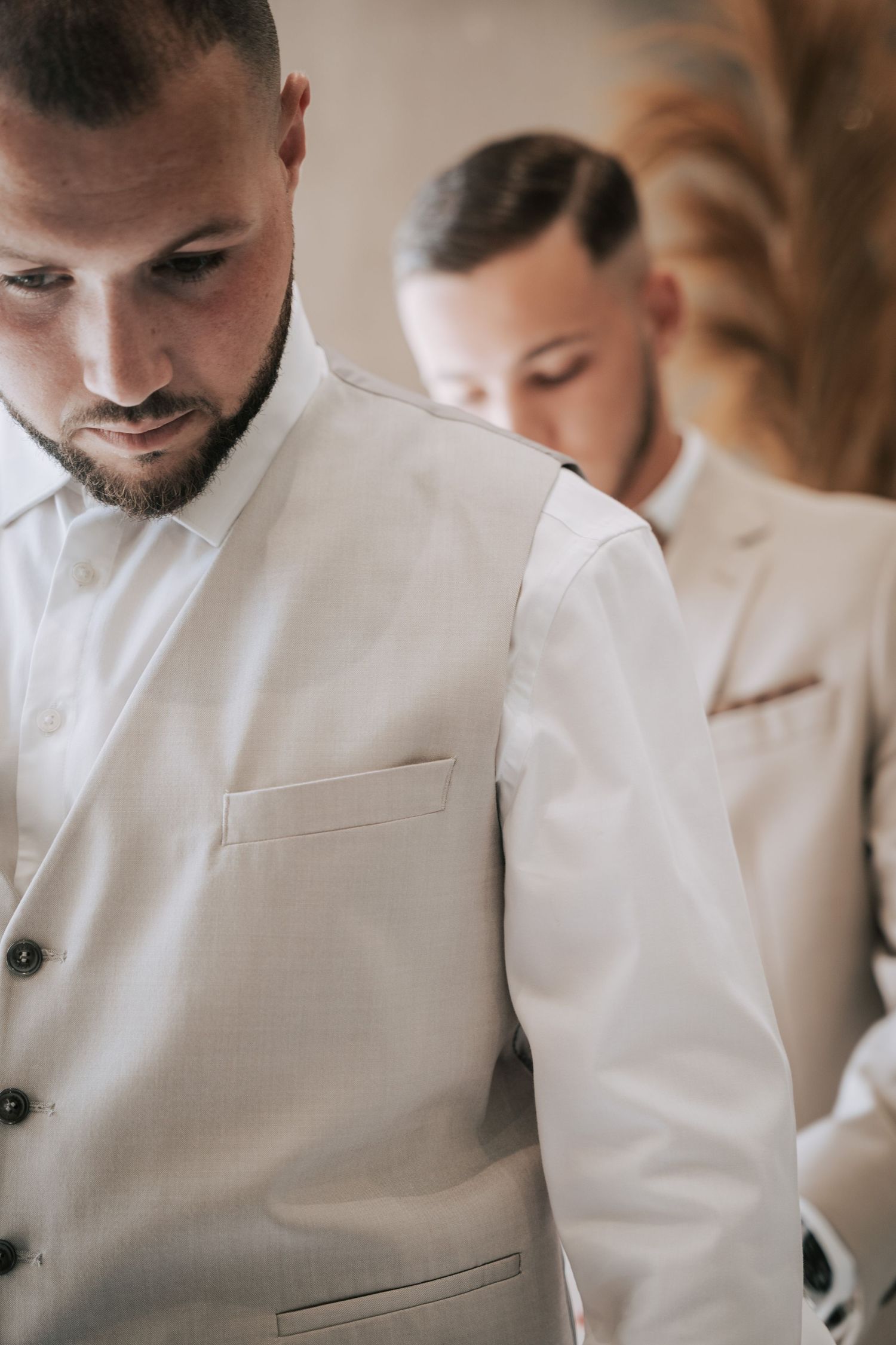Groom in white vest being helped by groomsman while getting ready for wedding ceremony.