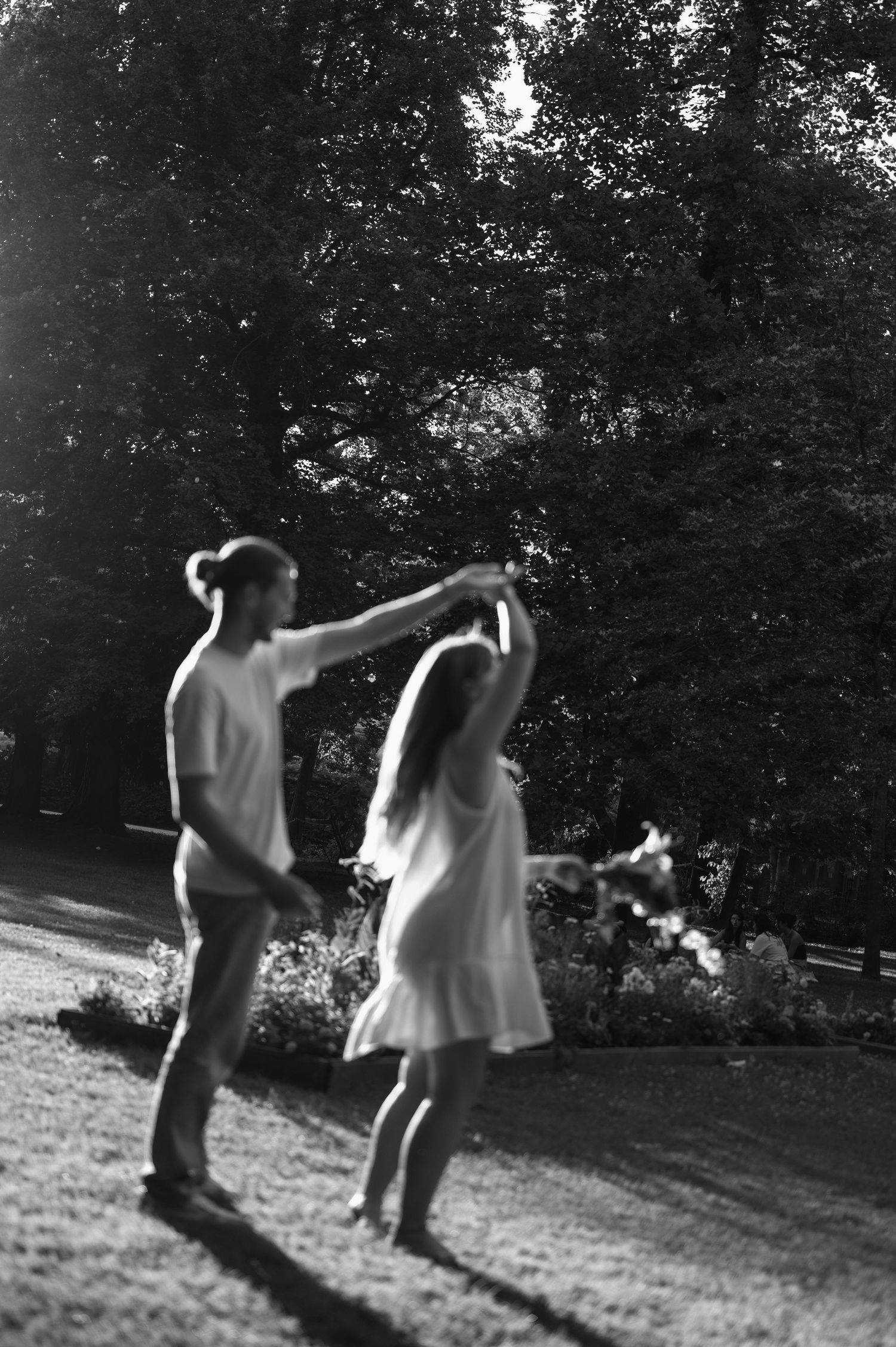 Black and white photo of a couple dancing together in a sunlit park surrounded by trees.
