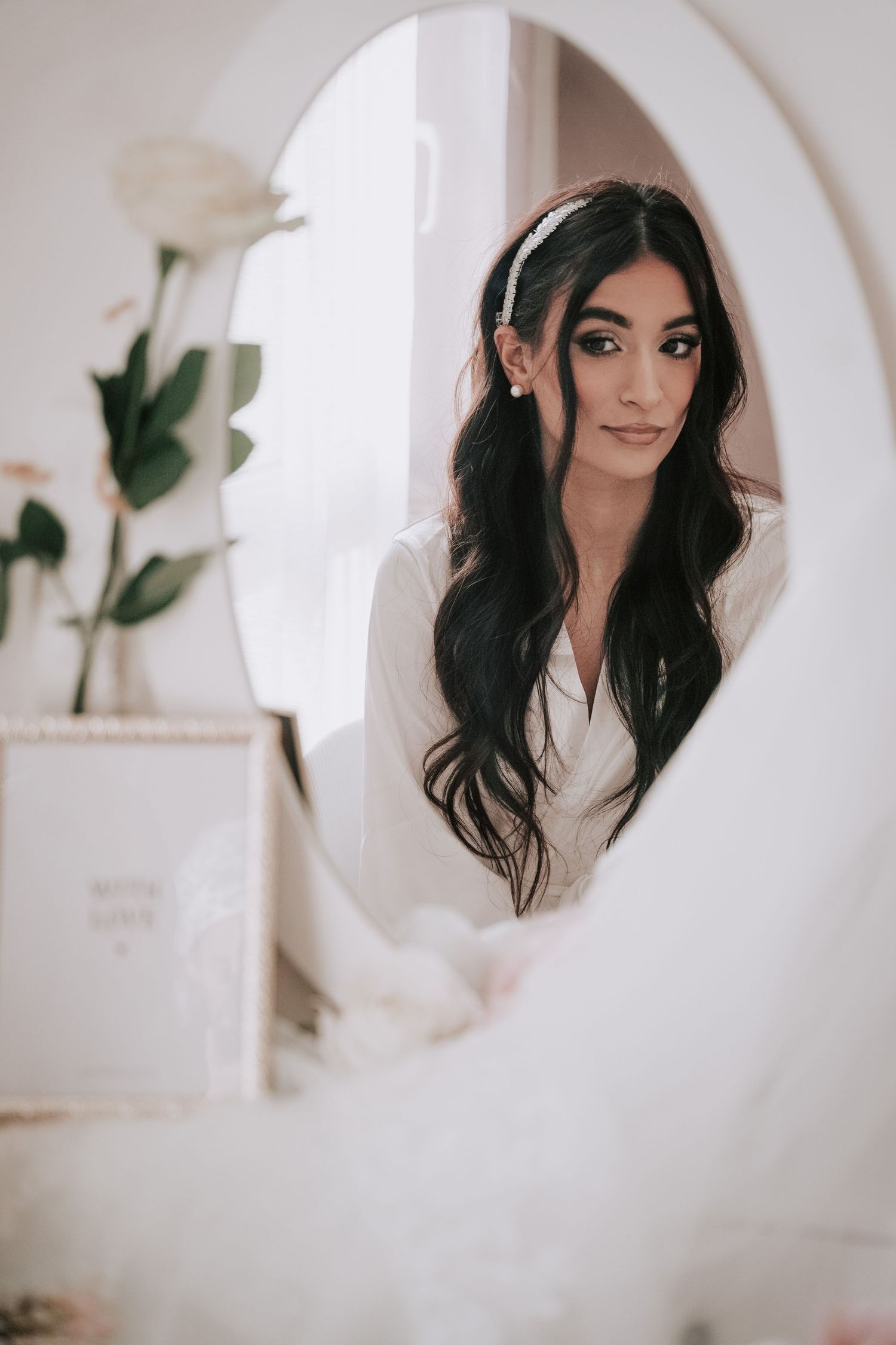 Beautiful dark-haired bride in white robe reflected in ornate oval mirror while getting ready.