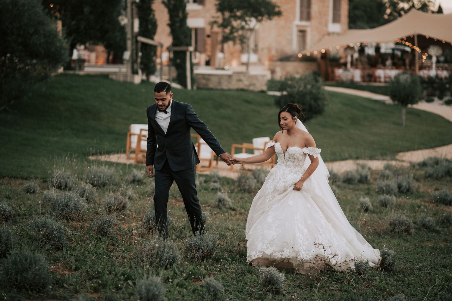 Joyful bride and groom stroll through lush garden grounds at elegant French countryside venue.