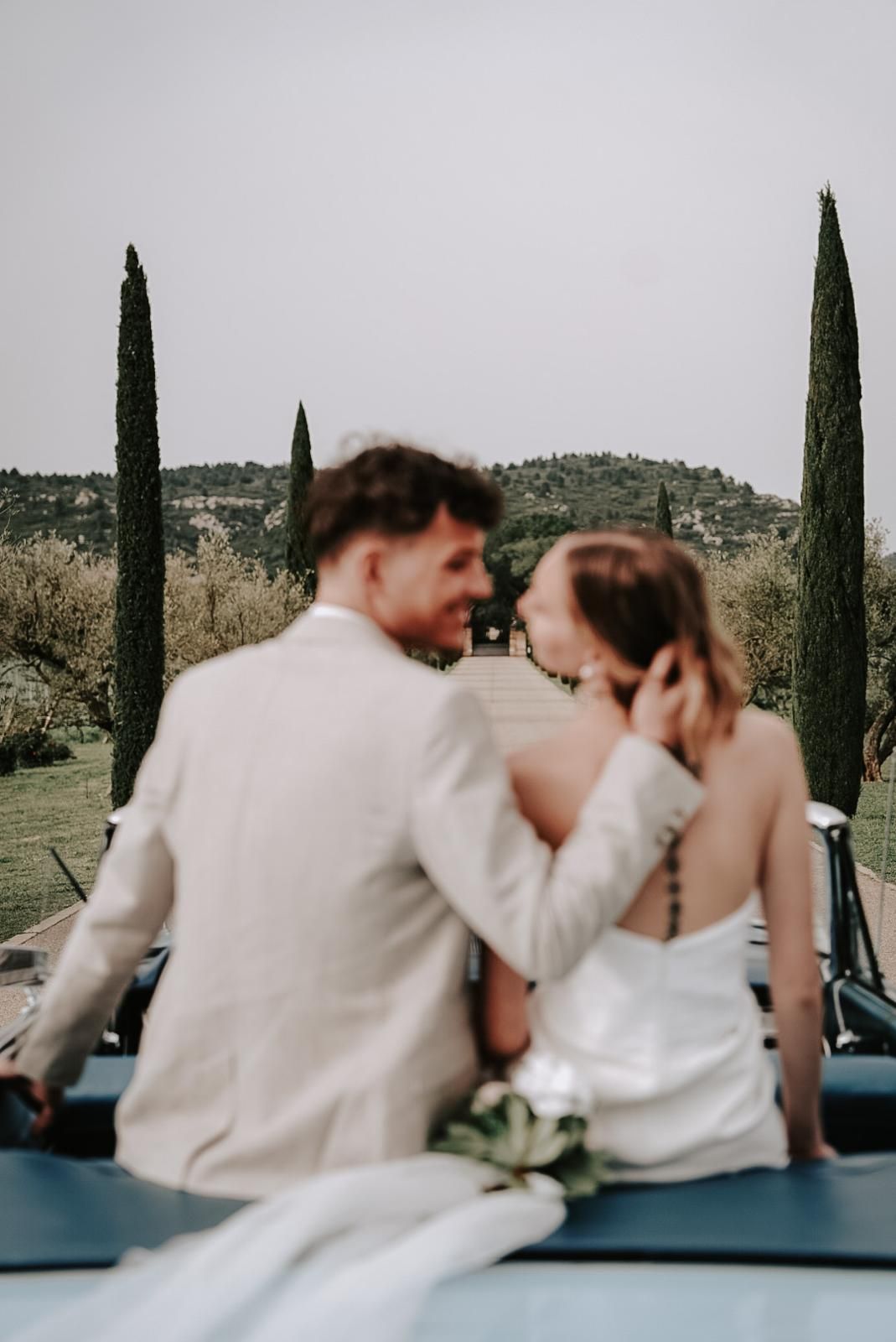 Bride and groom sit on a vintage blue convertible car, gazing at each other with cypress trees in the background.