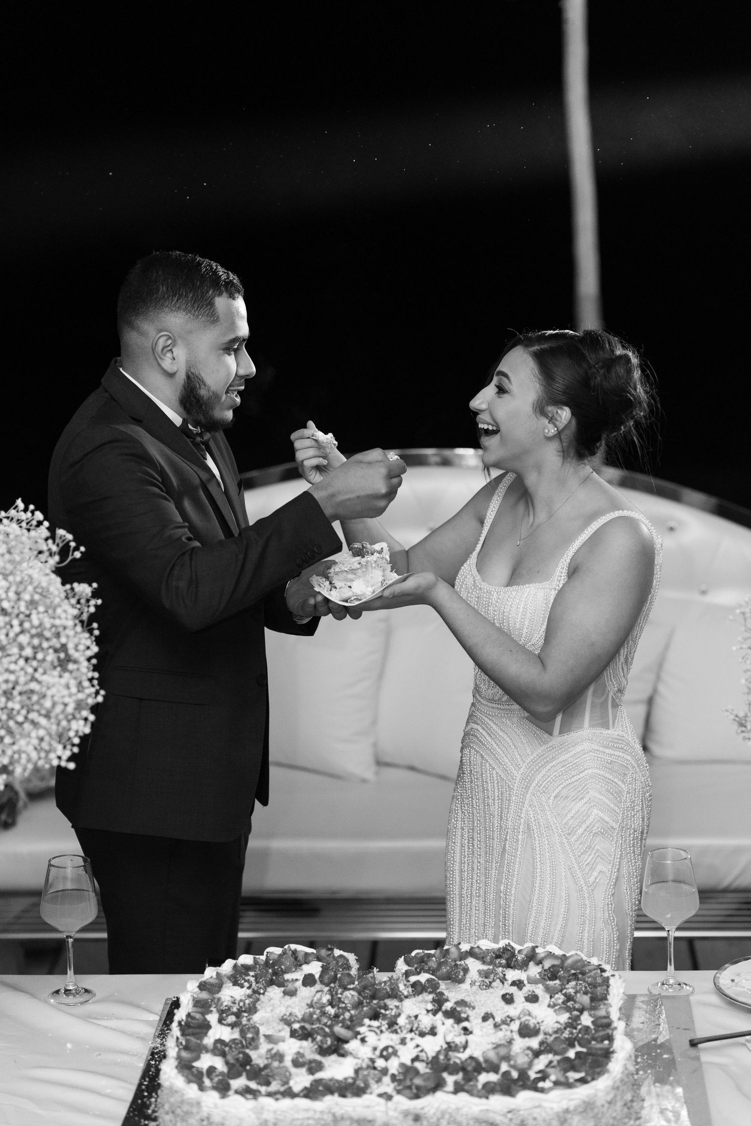 Black and white photo of groom feeding bride wedding cake slice at reception beside berry-topped cake.