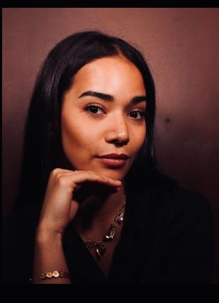 Young woman with long dark hair poses thoughtfully with hand under chin, wearing a necklace and bracelet.