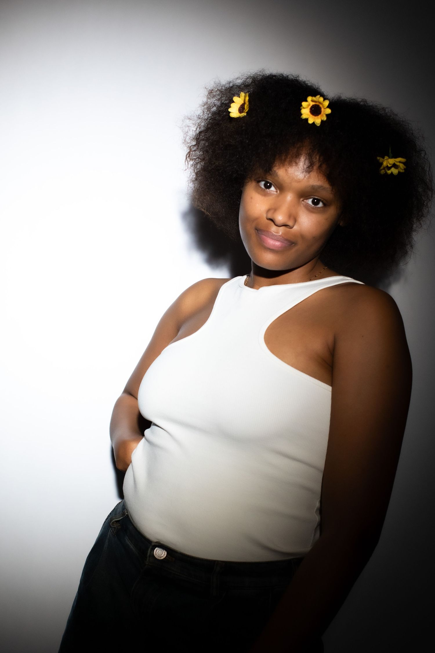 Young Black woman with curly afro adorned with yellow sunflowers, wearing white sleeveless top, posing confidently against gray background.