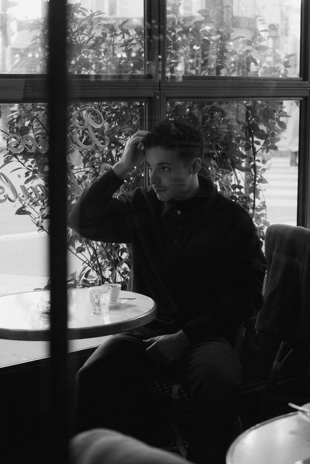 Black and white photo of a person sitting alone at a cafe table near a window, hand resting on their head.