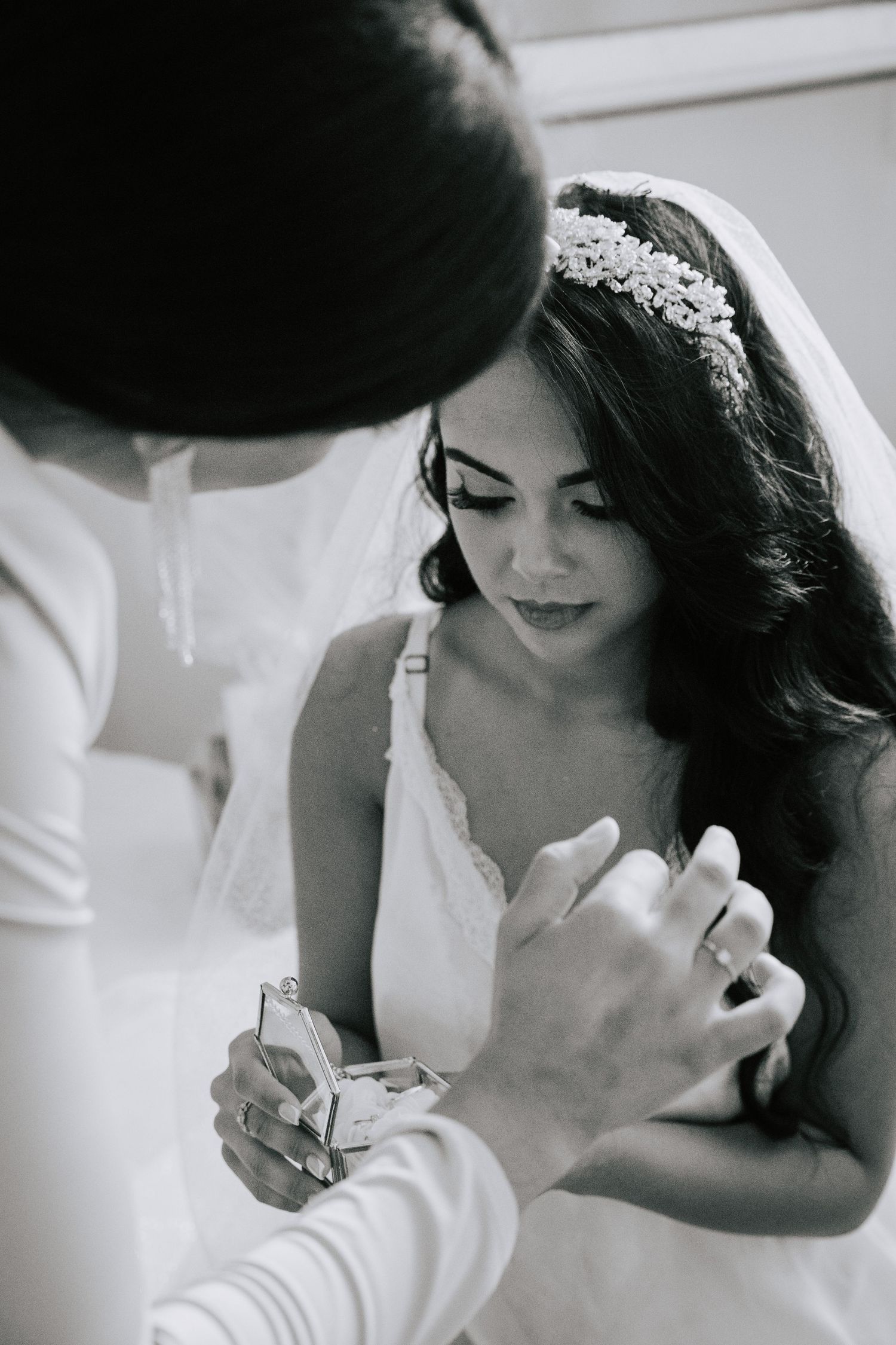 Black and white photo of a bride in lace gown and floral headpiece receiving a gift from an attendant.