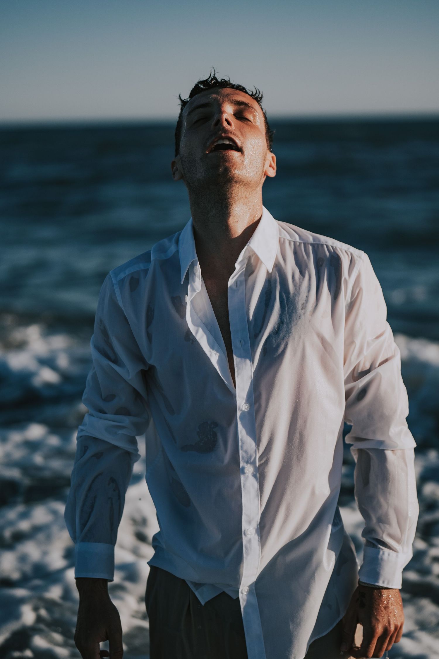 Man in wet white shirt standing at beach with head tilted back, ocean waves behind him.