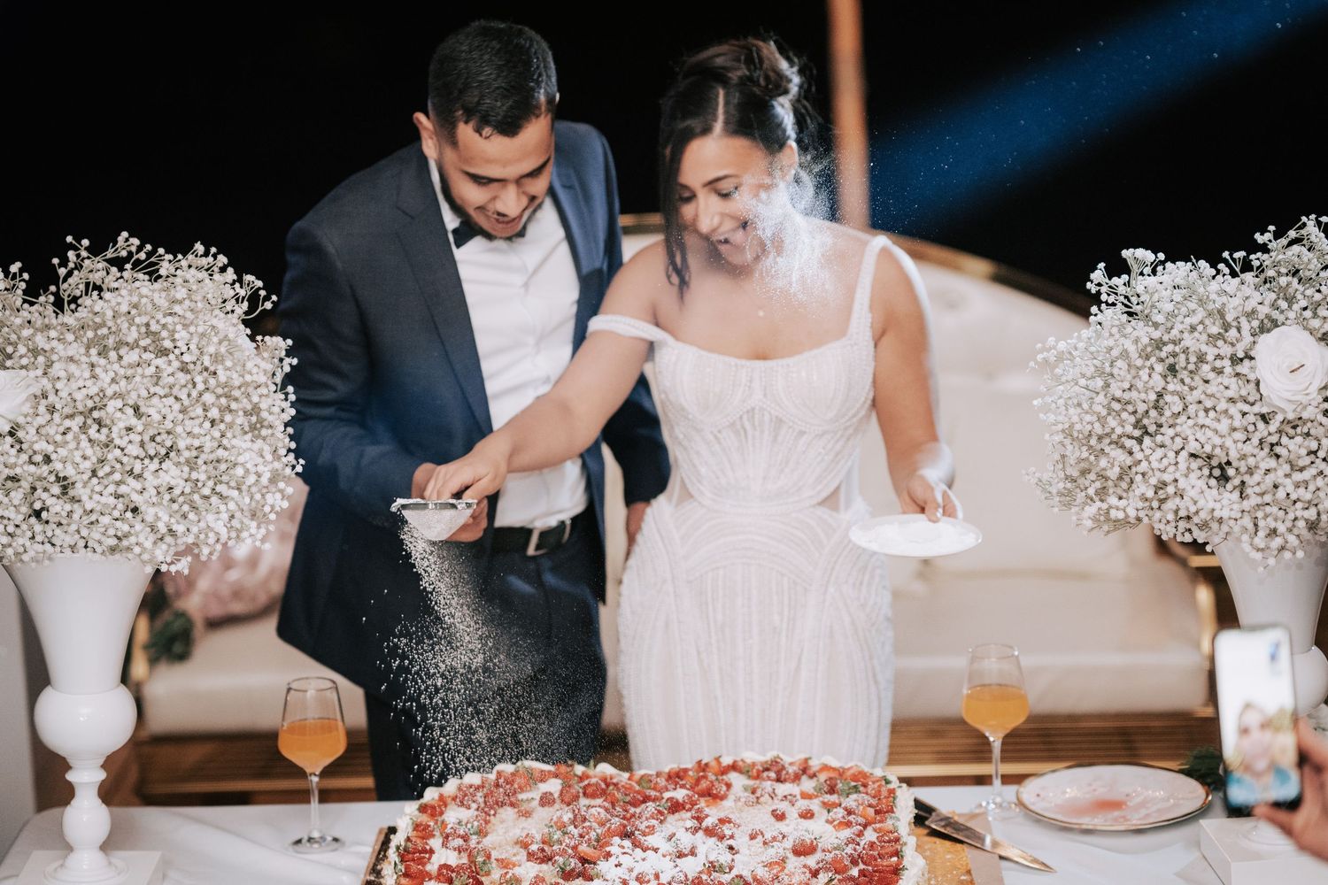 Bride and groom cutting wedding cake together as powdered sugar falls at outdoor evening reception celebration.