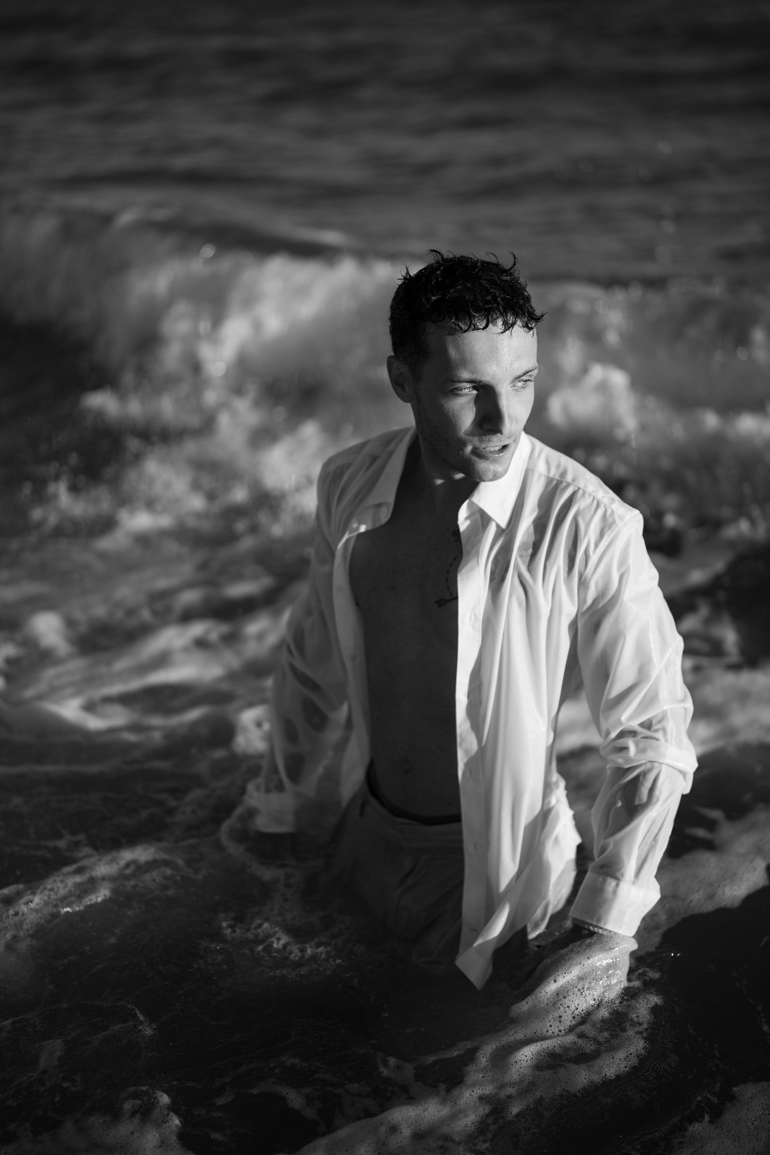 Man in soaked white shirt emerging from crashing ocean waves in dramatic black and white photo.