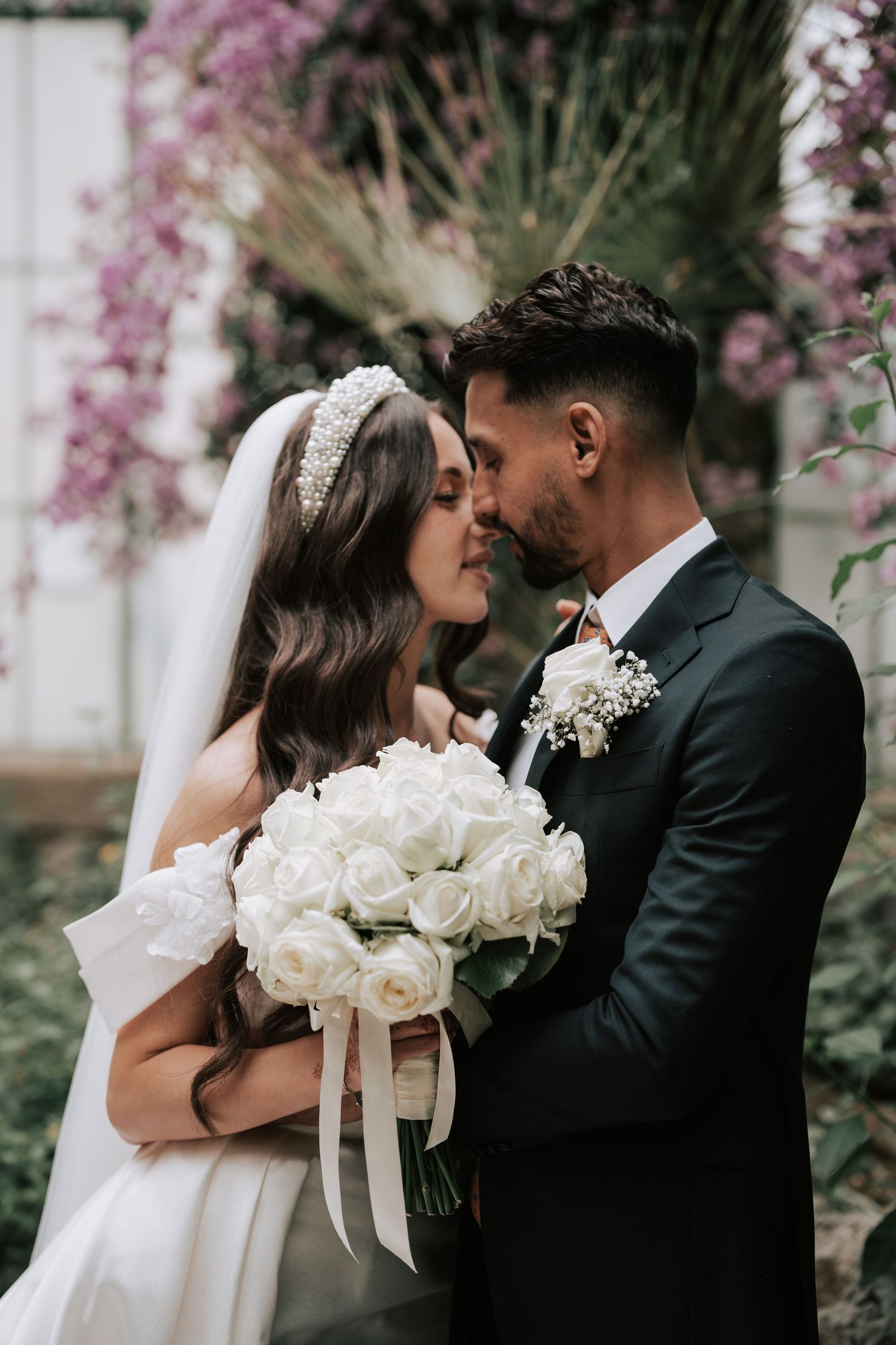 Bride and groom sharing a romantic almost-kiss holding white rose bouquet with purple floral backdrop.