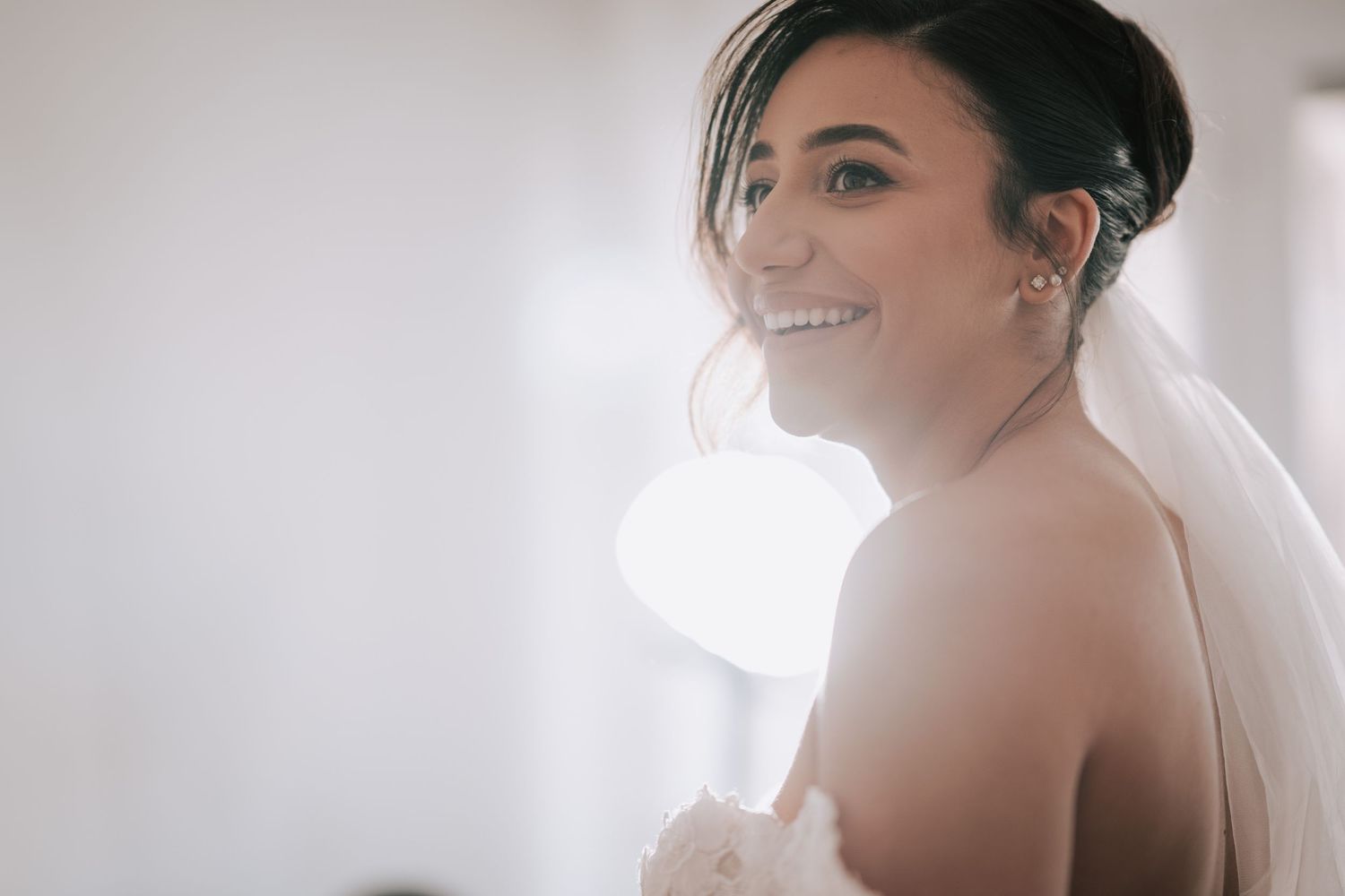 Radiant bride in a veil smiles softly in bright natural light on her wedding day.