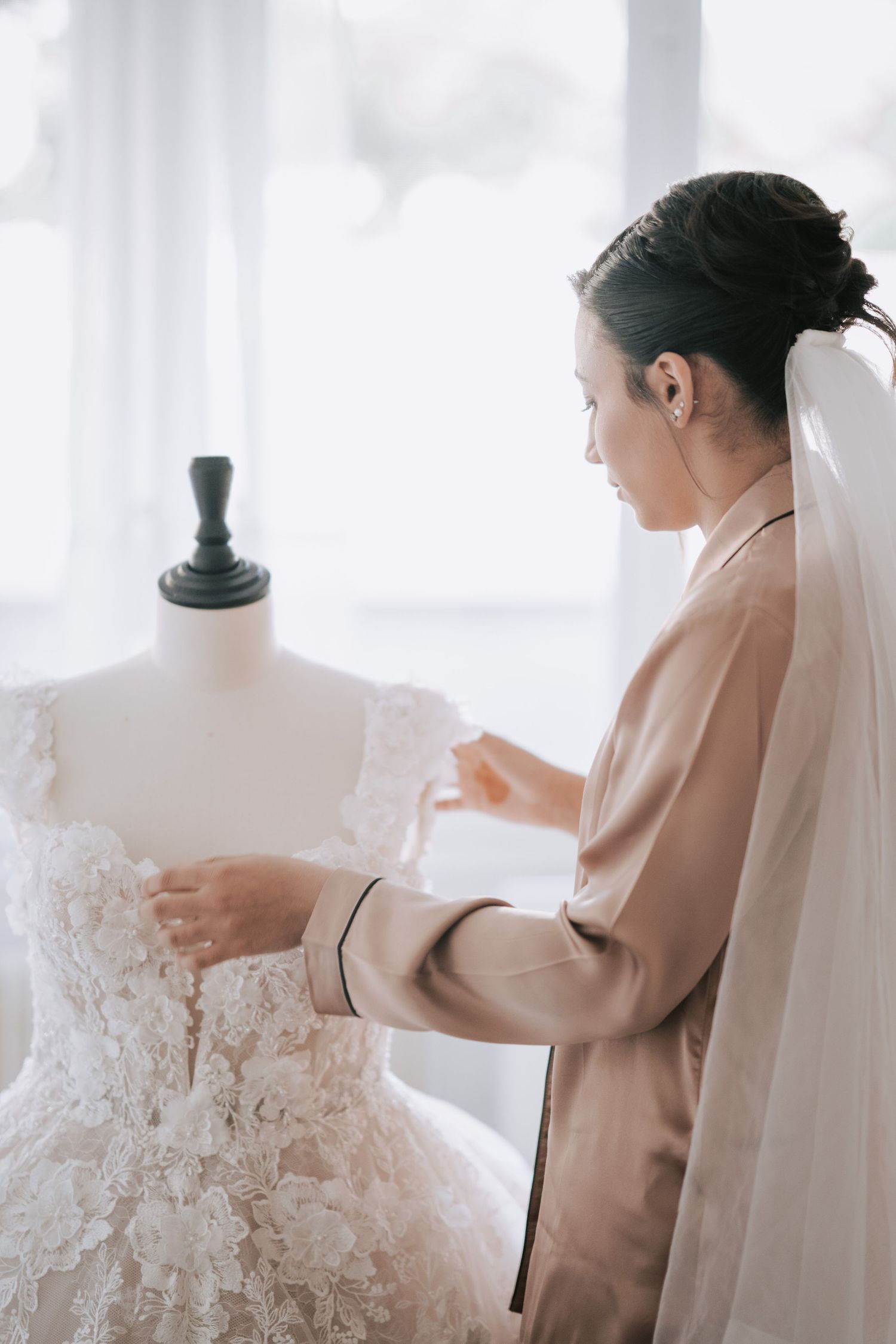 Bride in silk robe adjusting floral lace wedding dress on mannequin with veil.