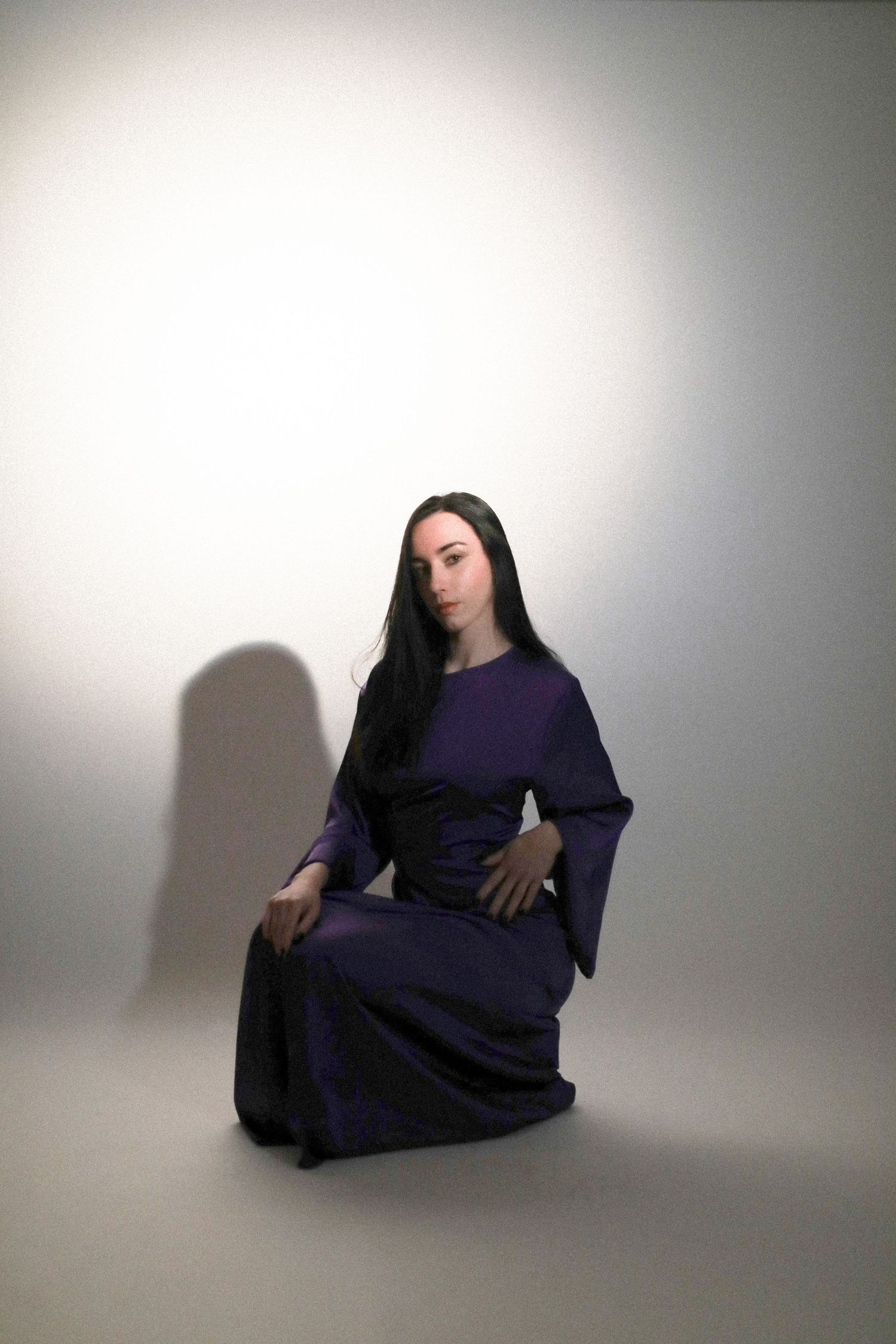 Woman in purple medieval gown with long dark hair seated gracefully against a neutral gray background.