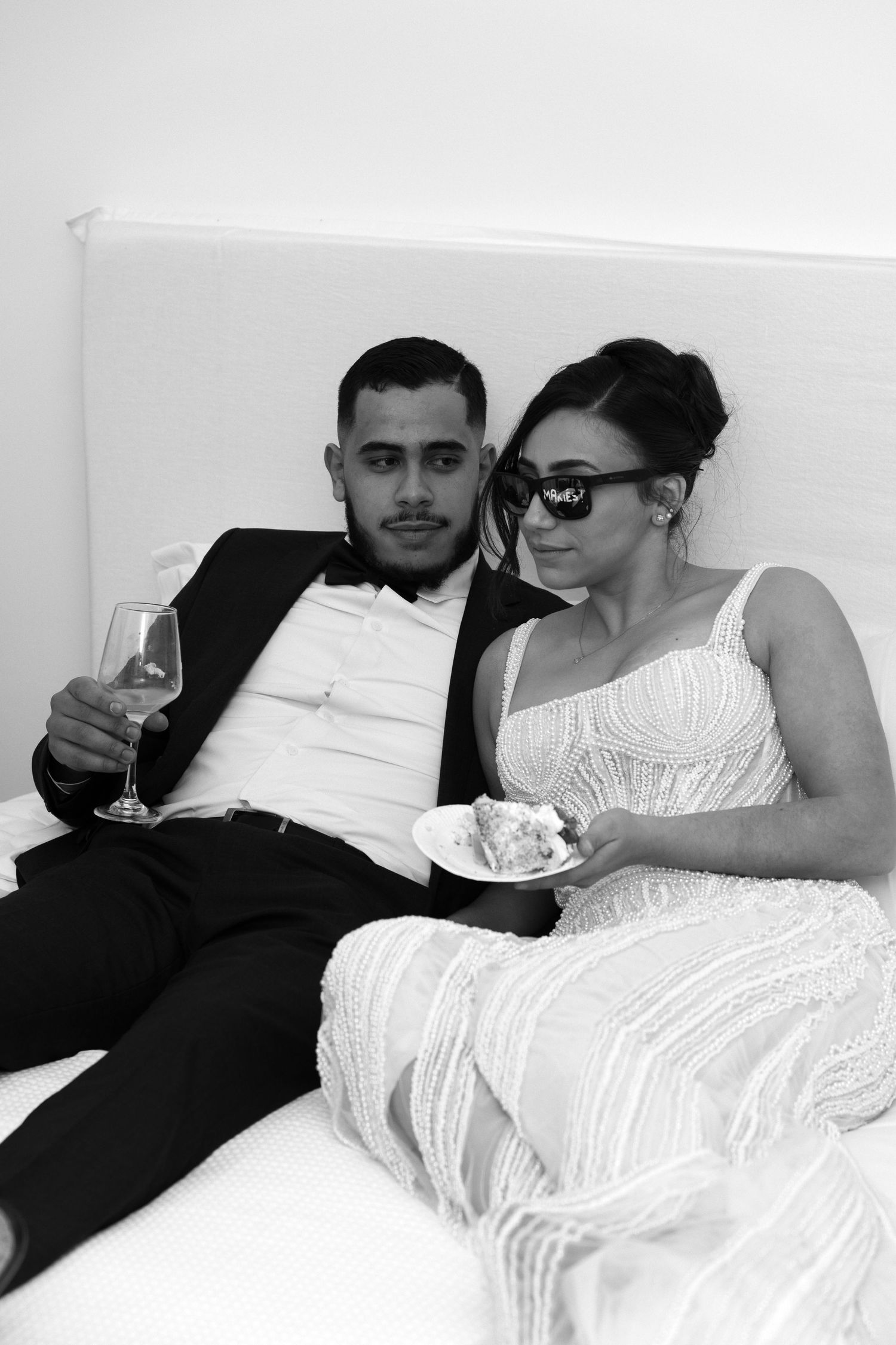Bride and groom relaxing on a bed in black and white, groom holding champagne glass, bride wearing sunglasses with cake.