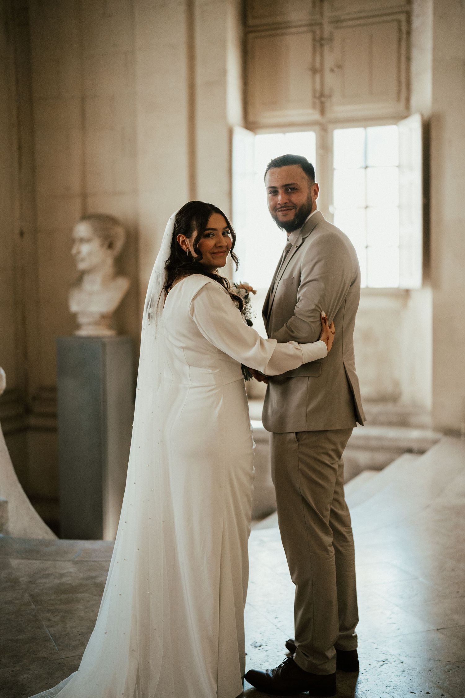 Bride and groom embracing and smiling inside a classical hall with marble busts on their wedding day.