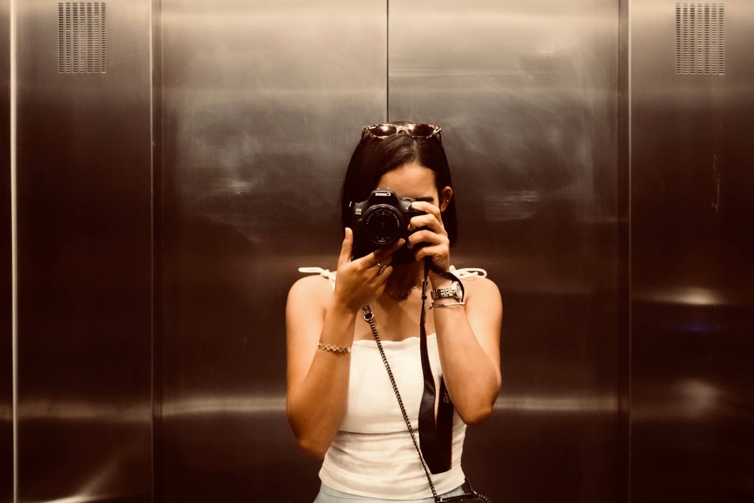 Woman taking a self-portrait with a DSLR camera in a reflective metal elevator.