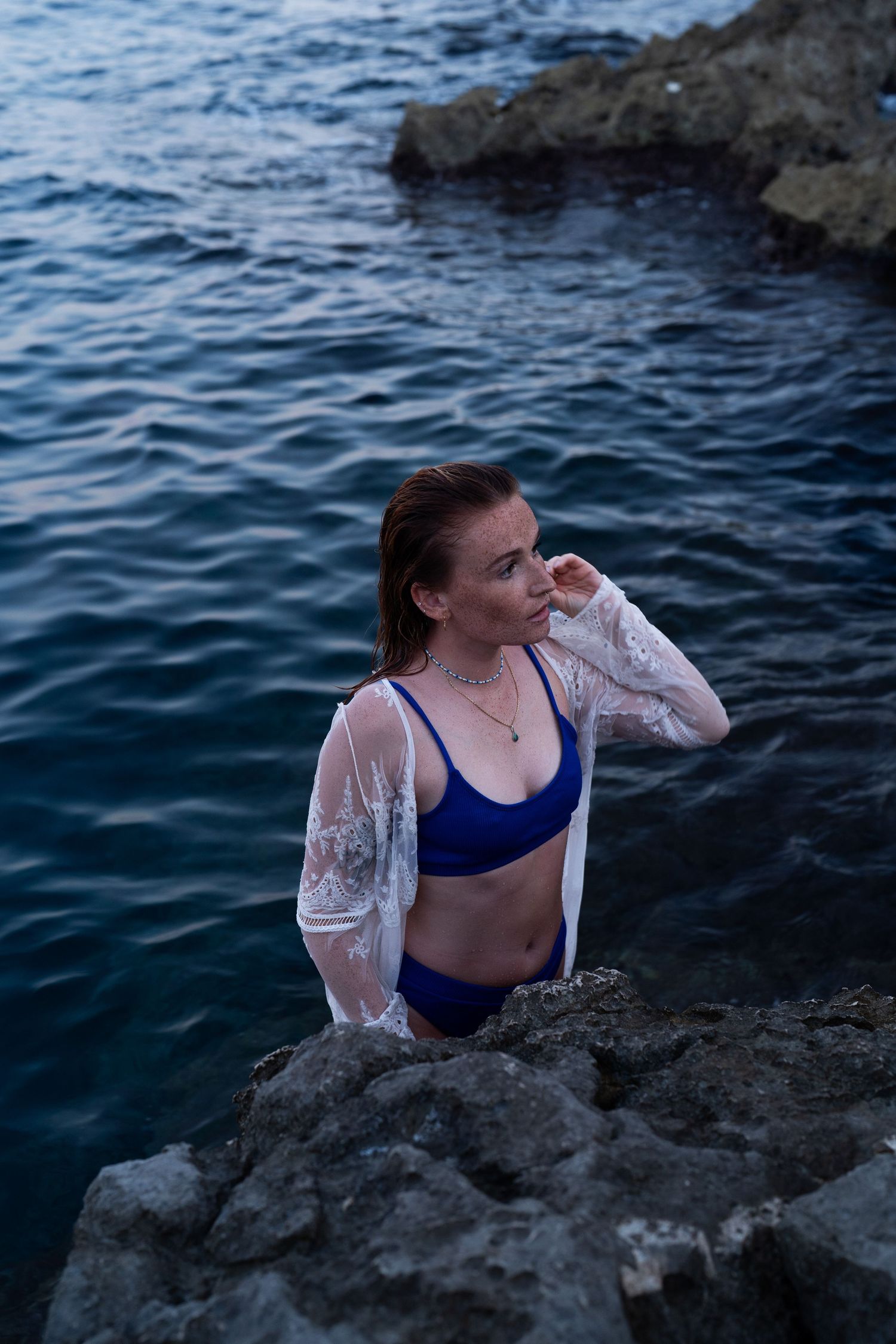 Woman in blue bikini set and white lace cardigan standing among rocks in calm ocean water.