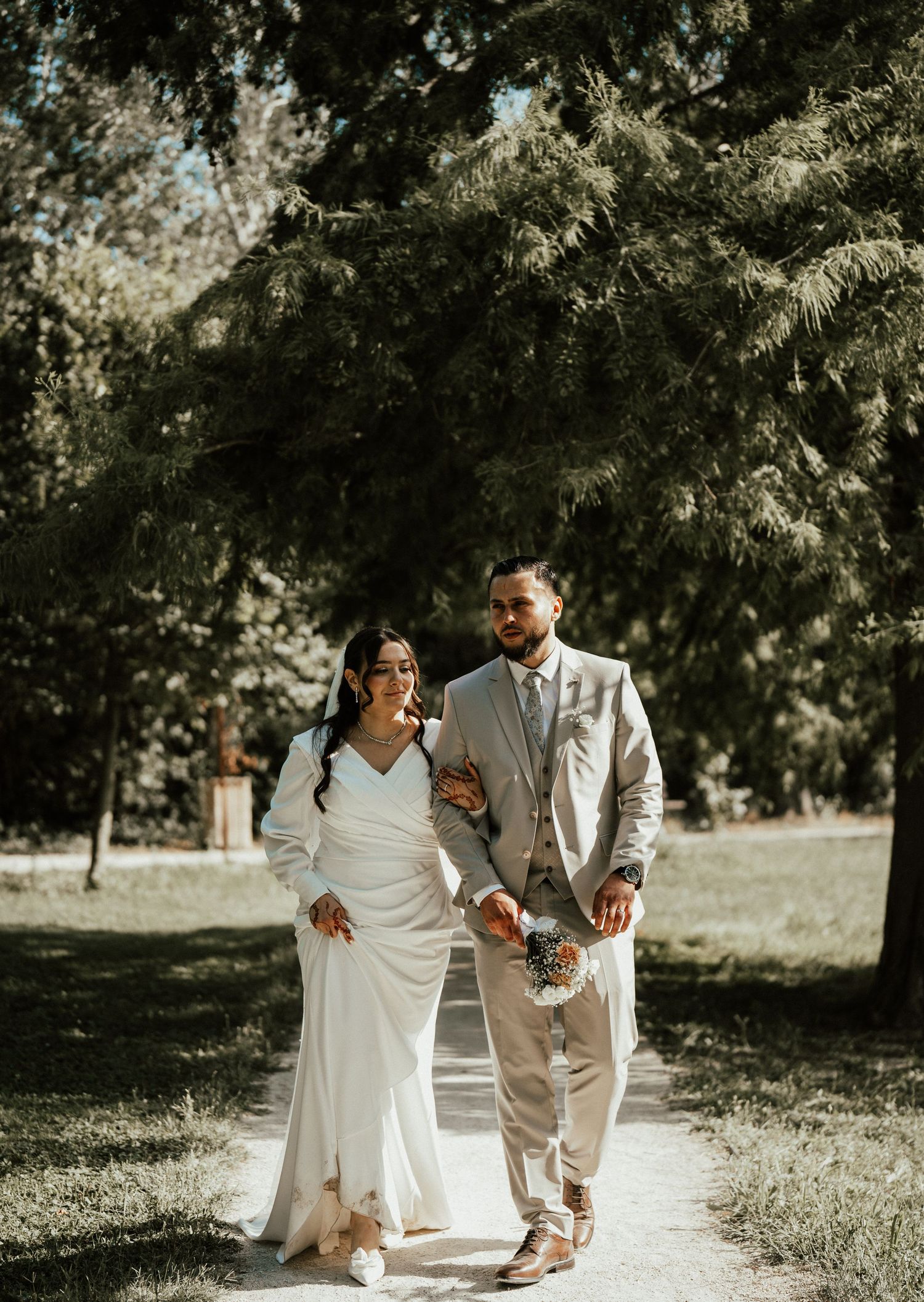 Bride and groom walking together along a sunlit garden path surrounded by tall trees on wedding day.