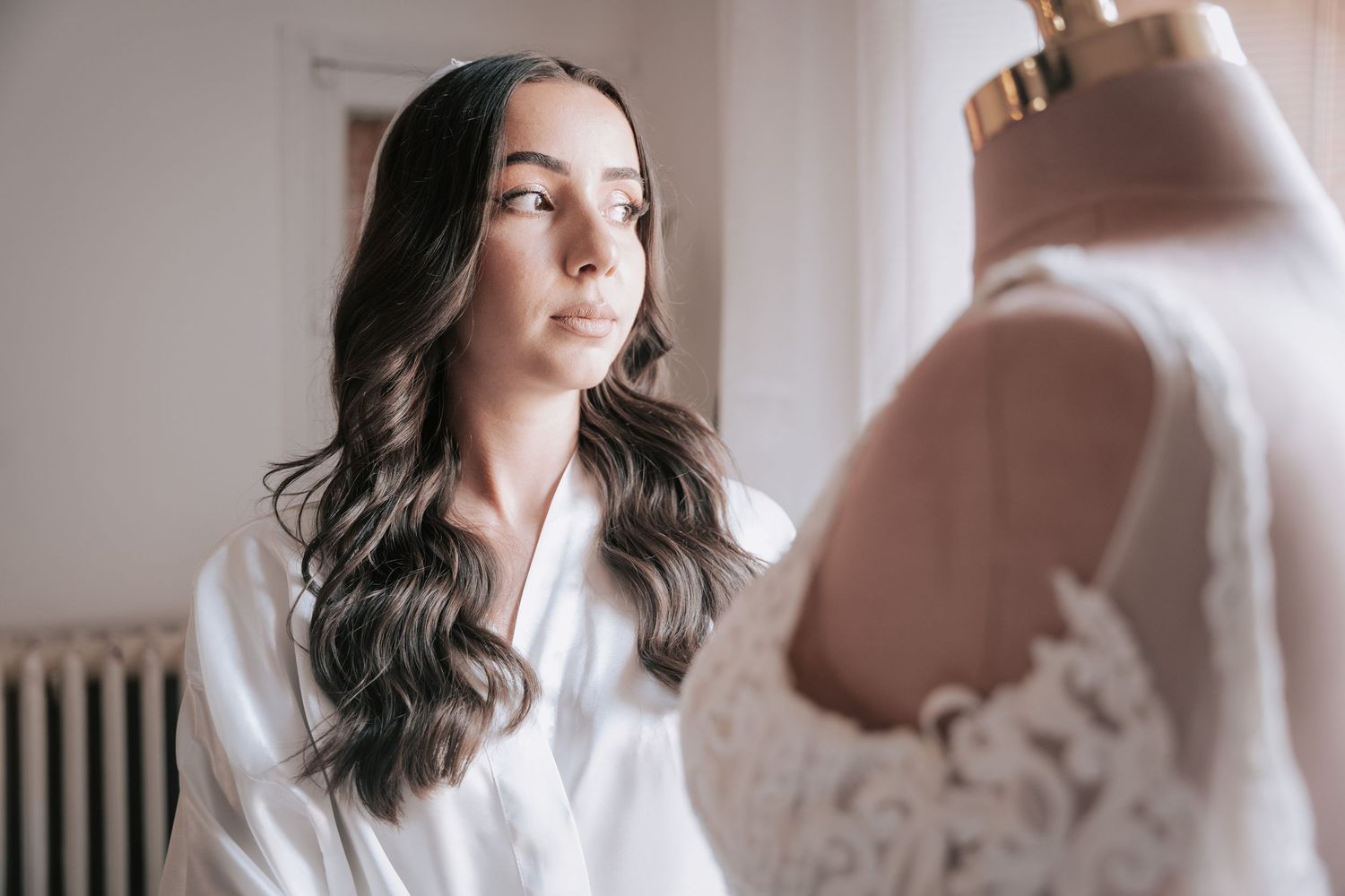 Brunette bride in white robe gazing at lace wedding gown on dress form mannequin.