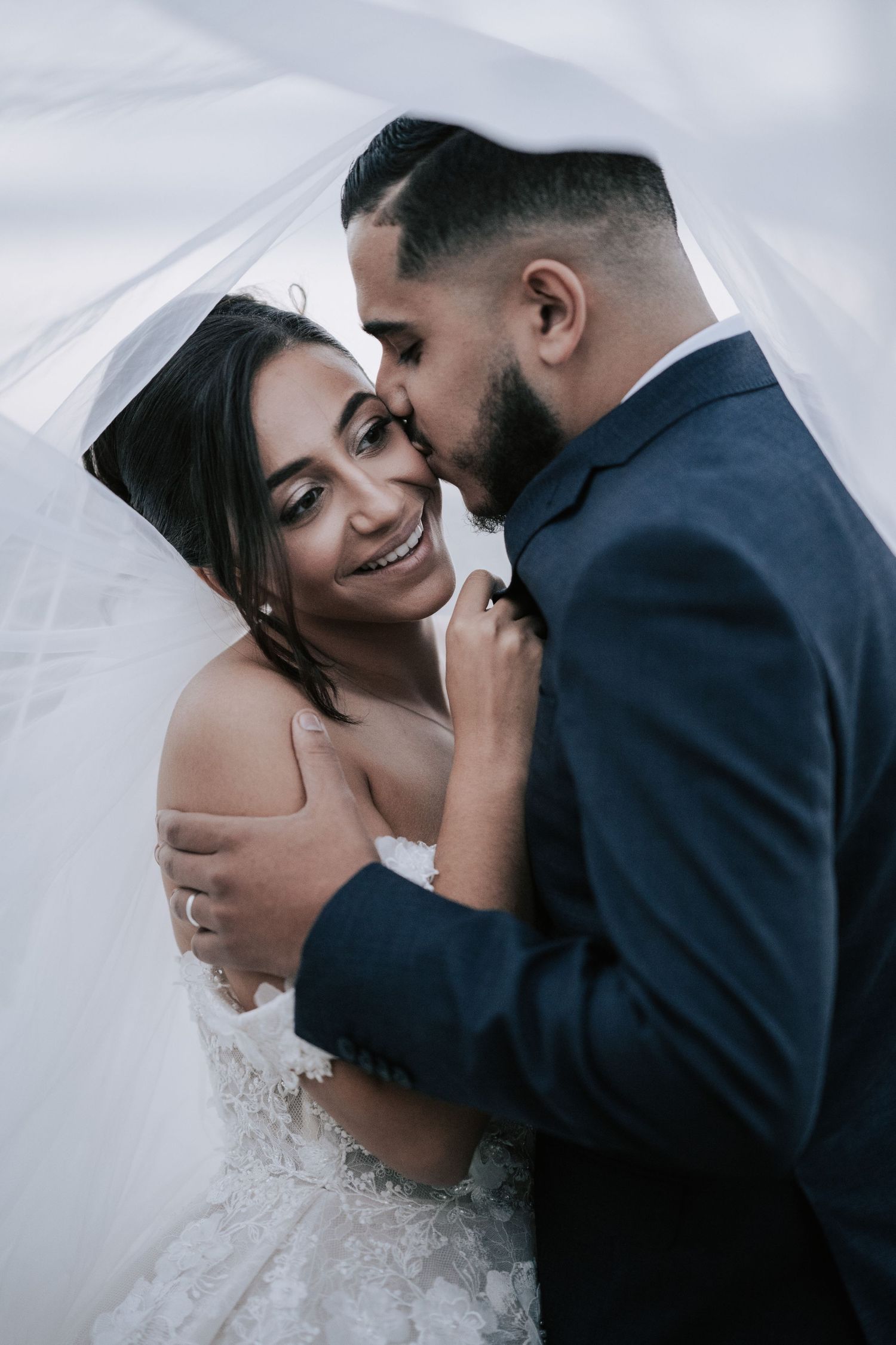 Groom kisses brides cheek as she smiles joyfully under a flowing veil in a romantic wedding portrait.