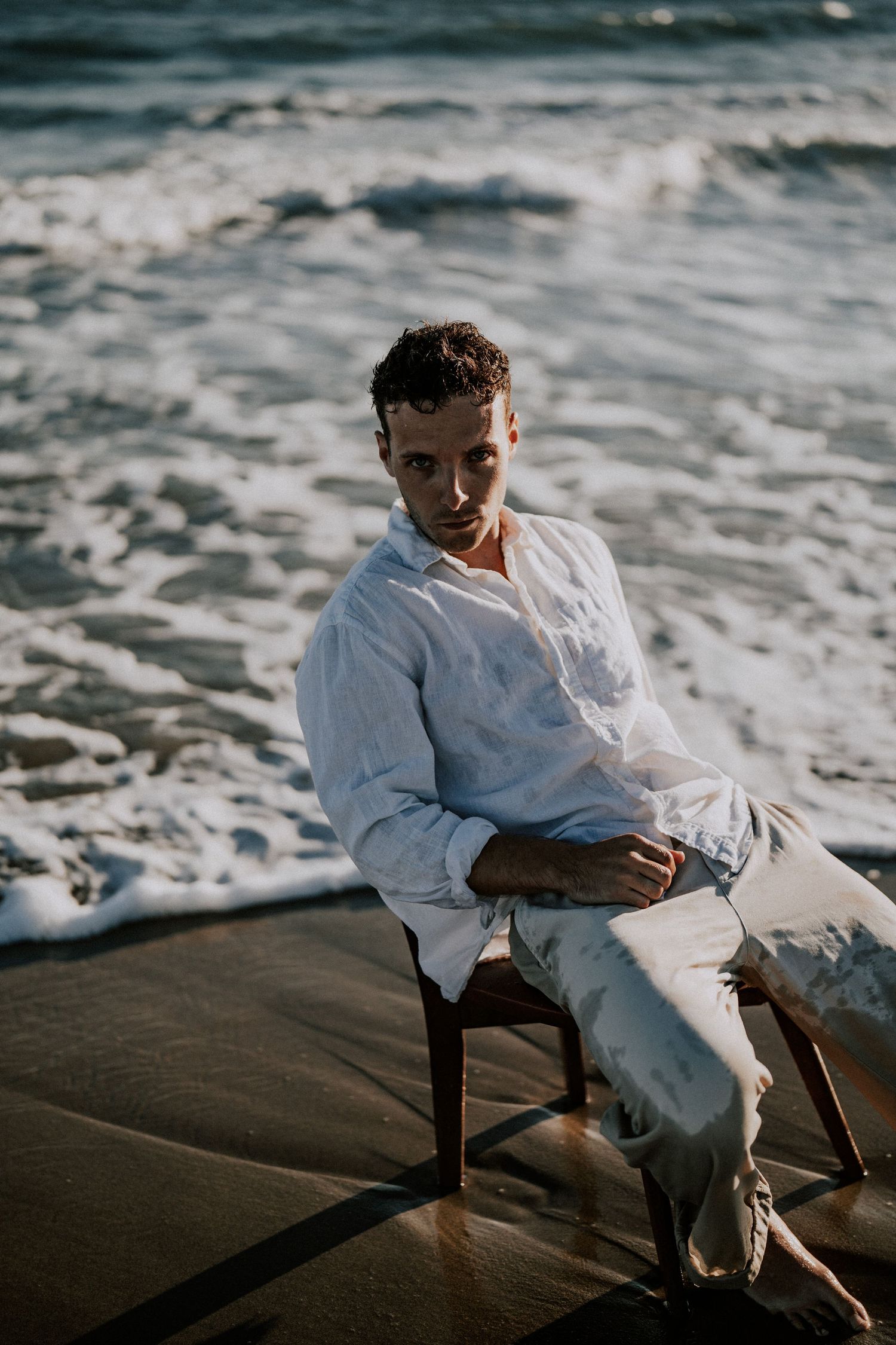A young man in a white linen shirt sits on a wooden chair on a sandy beach with ocean waves.