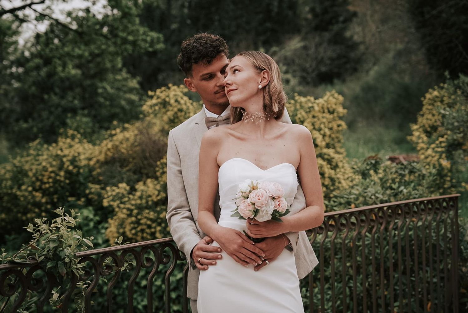 Bride in strapless white gown holds pink bouquet as groom in beige suit embraces her outdoors.